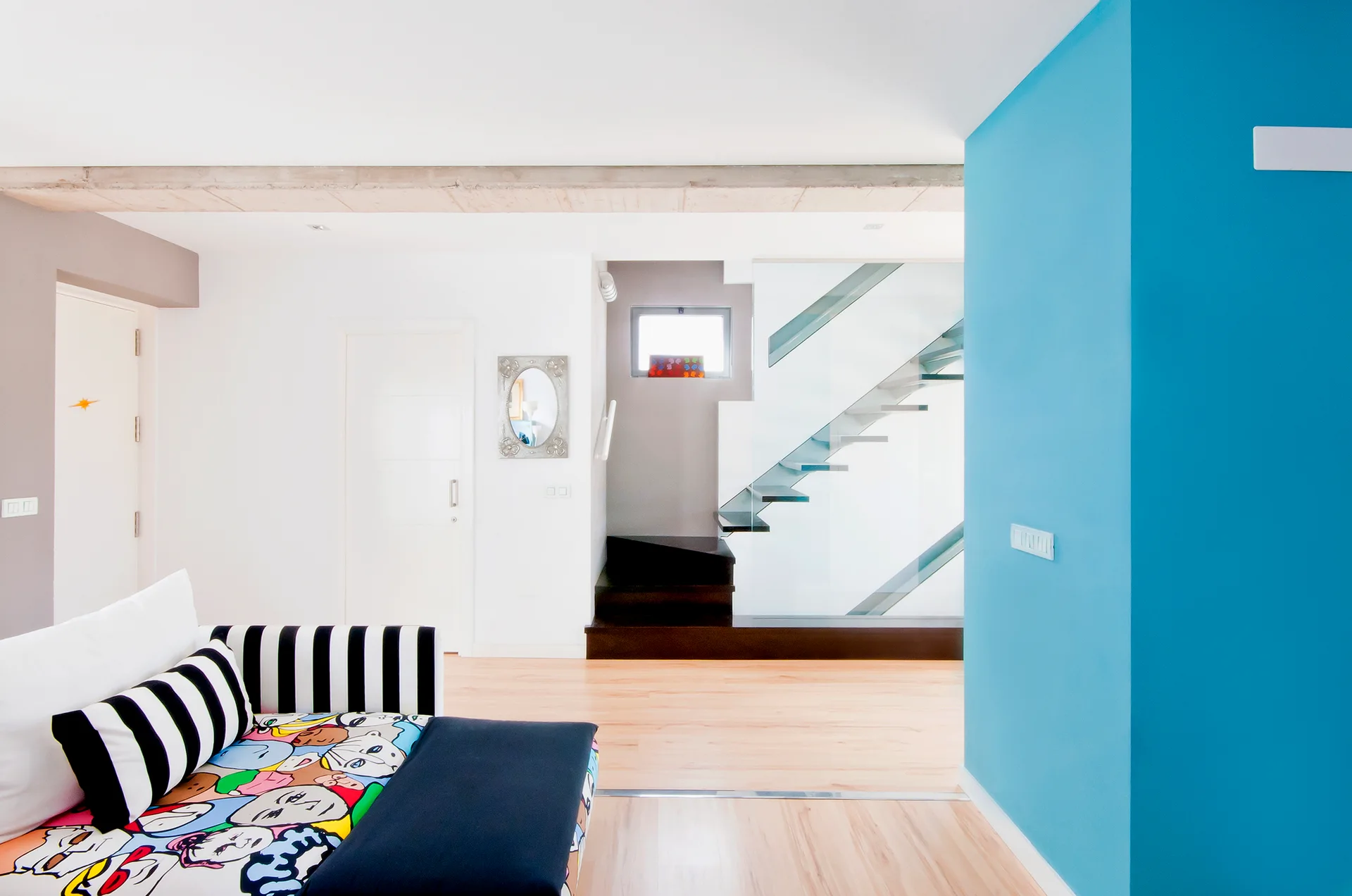 A modern living space features a graphic cartoon-print sofa with black and white stripes, a bright blue accent wall, and a floating glass staircase.