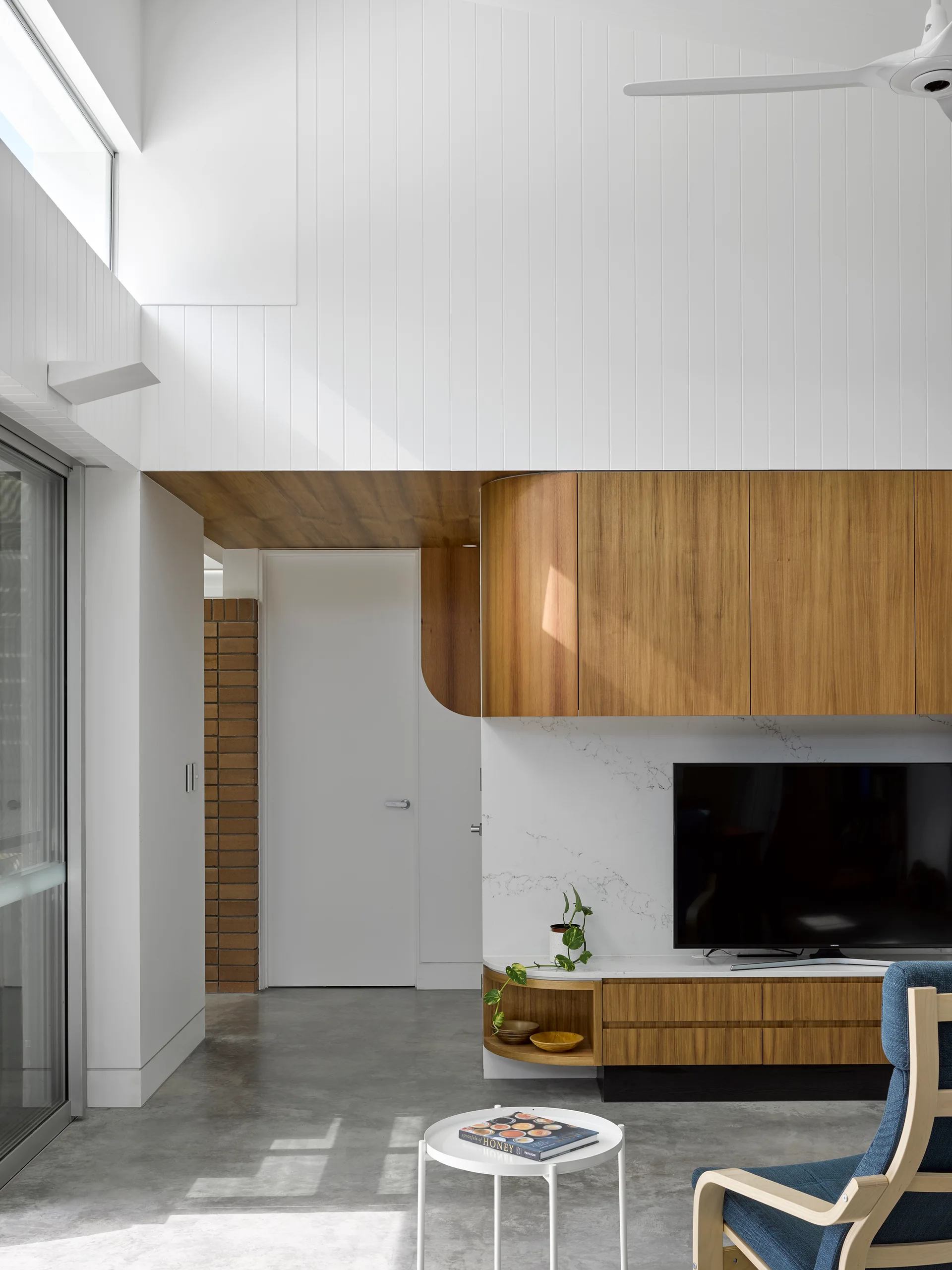 A modern living room features a custom curved wood media unit, a white paneled wall, polished concrete flooring, and a blue armchair.