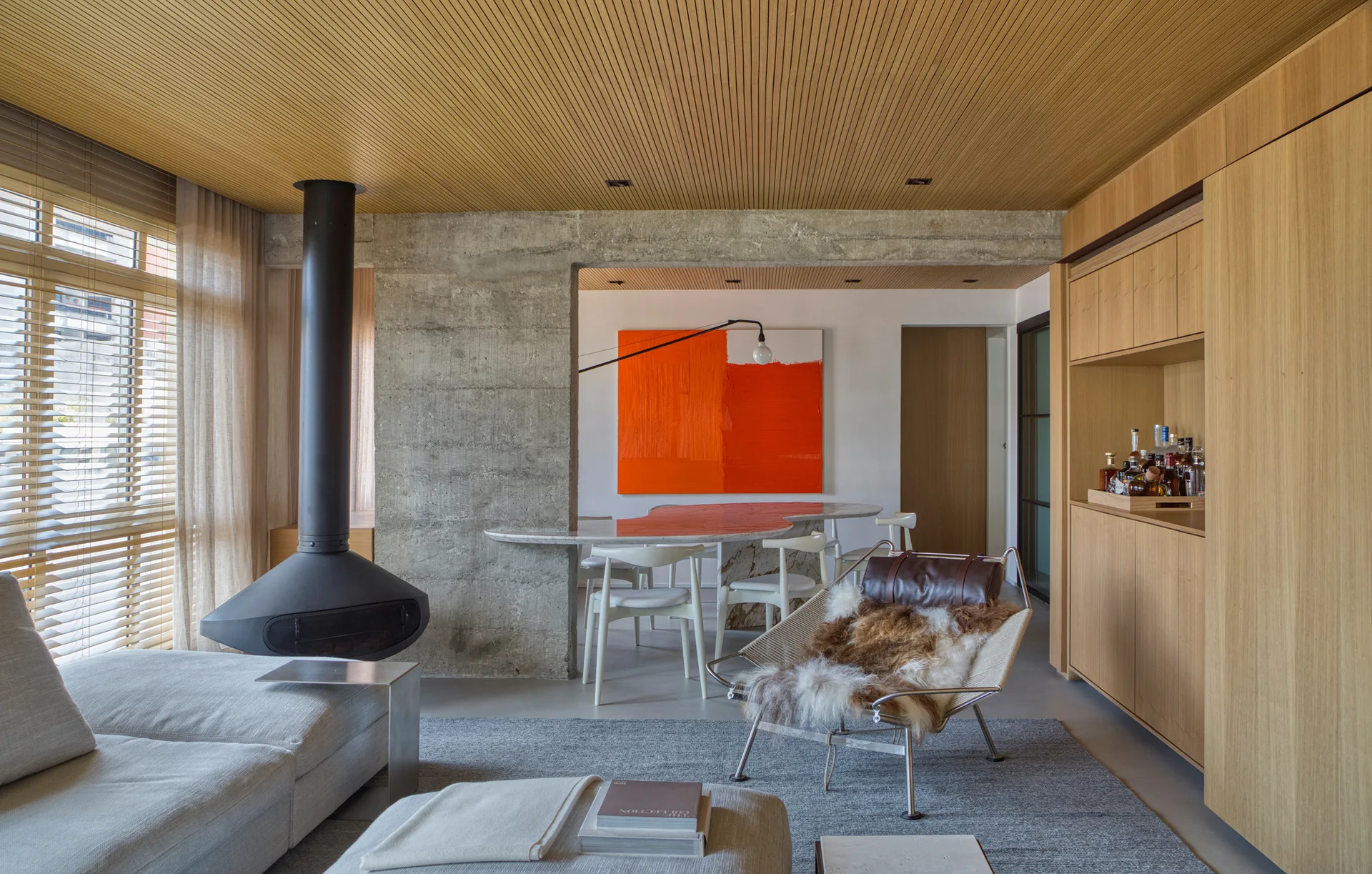 A modern living room features a hanging black fireplace, a textured concrete pillar, slatted wood ceilings, and a gray sofa on a textured rug.