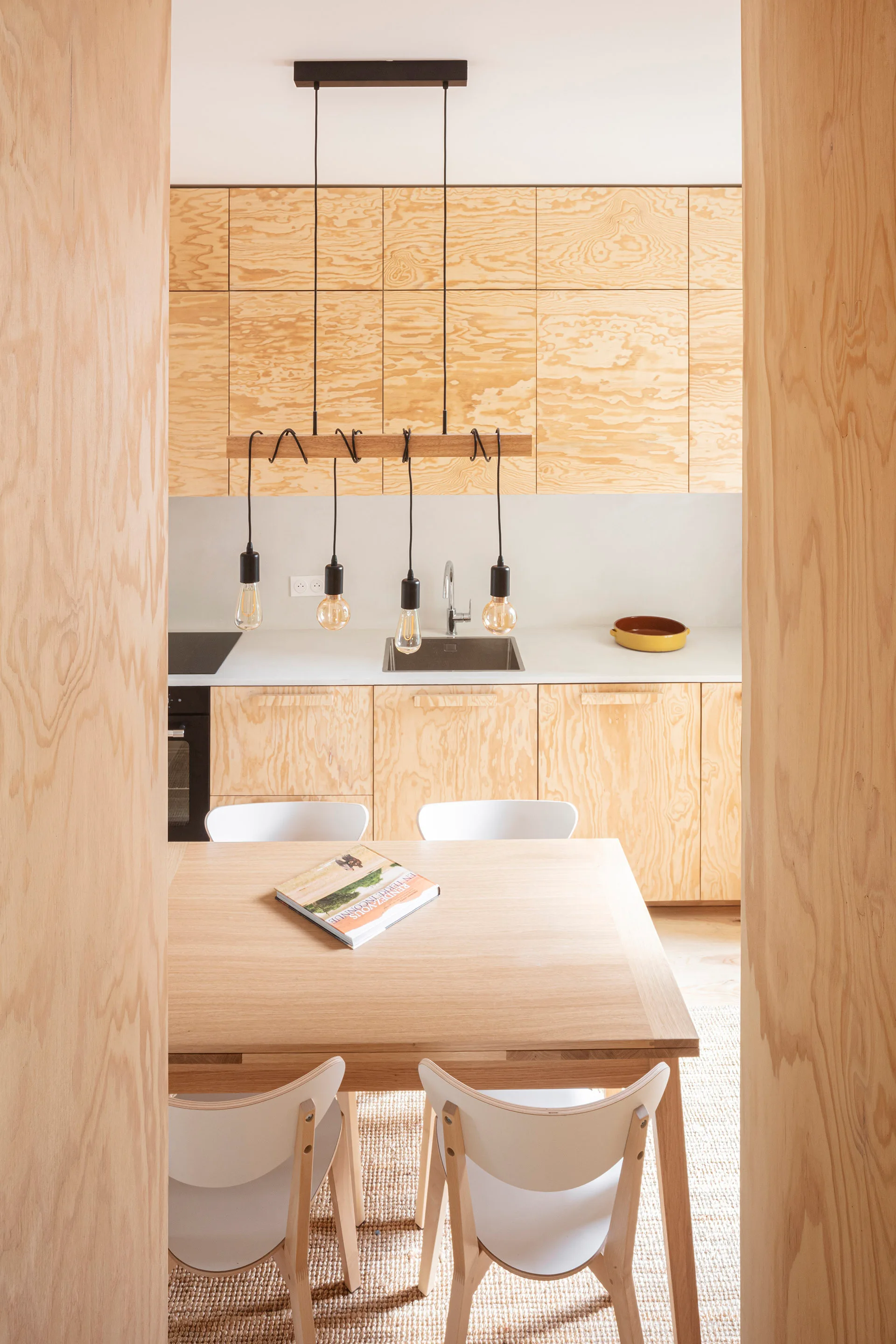 A dining table with white chairs sits in front of kitchen cabinets made of light-colored plywood with prominent wood grain.