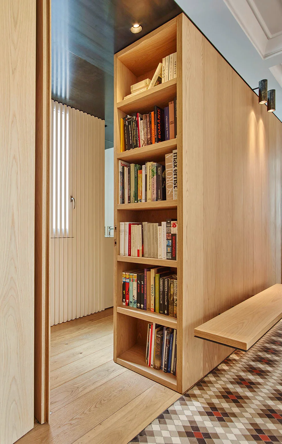 A light wood built-in bookshelf serves as a partition in a hallway next to a floating bench, wood paneling, and geometric tile flooring.