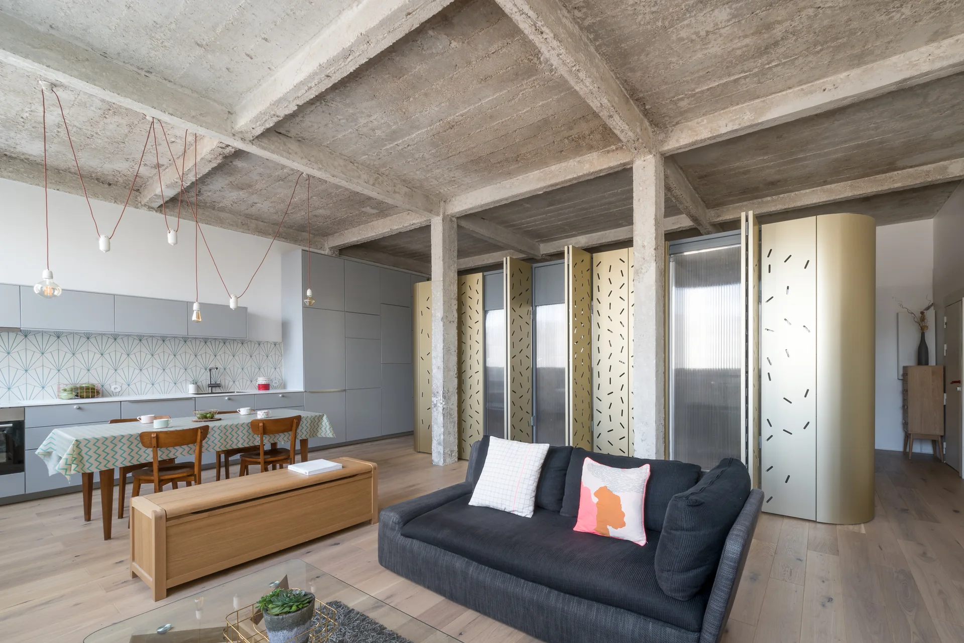 An open-concept living area and grey kitchen featuring an exposed concrete ceiling, a charcoal sofa, and patterned gold folding screens.