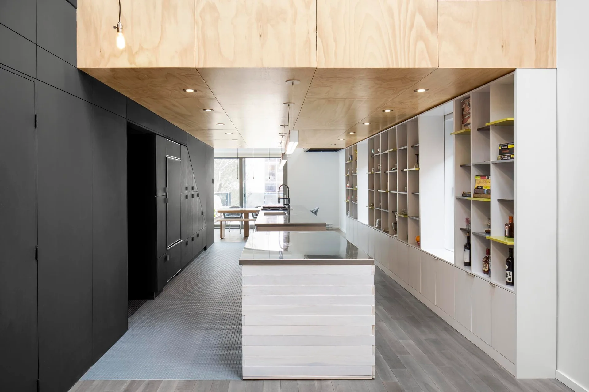 A long modern kitchen features a white-washed wood island, full-height matte black cabinets, and a natural plywood ceiling soffit with recessed lights.
