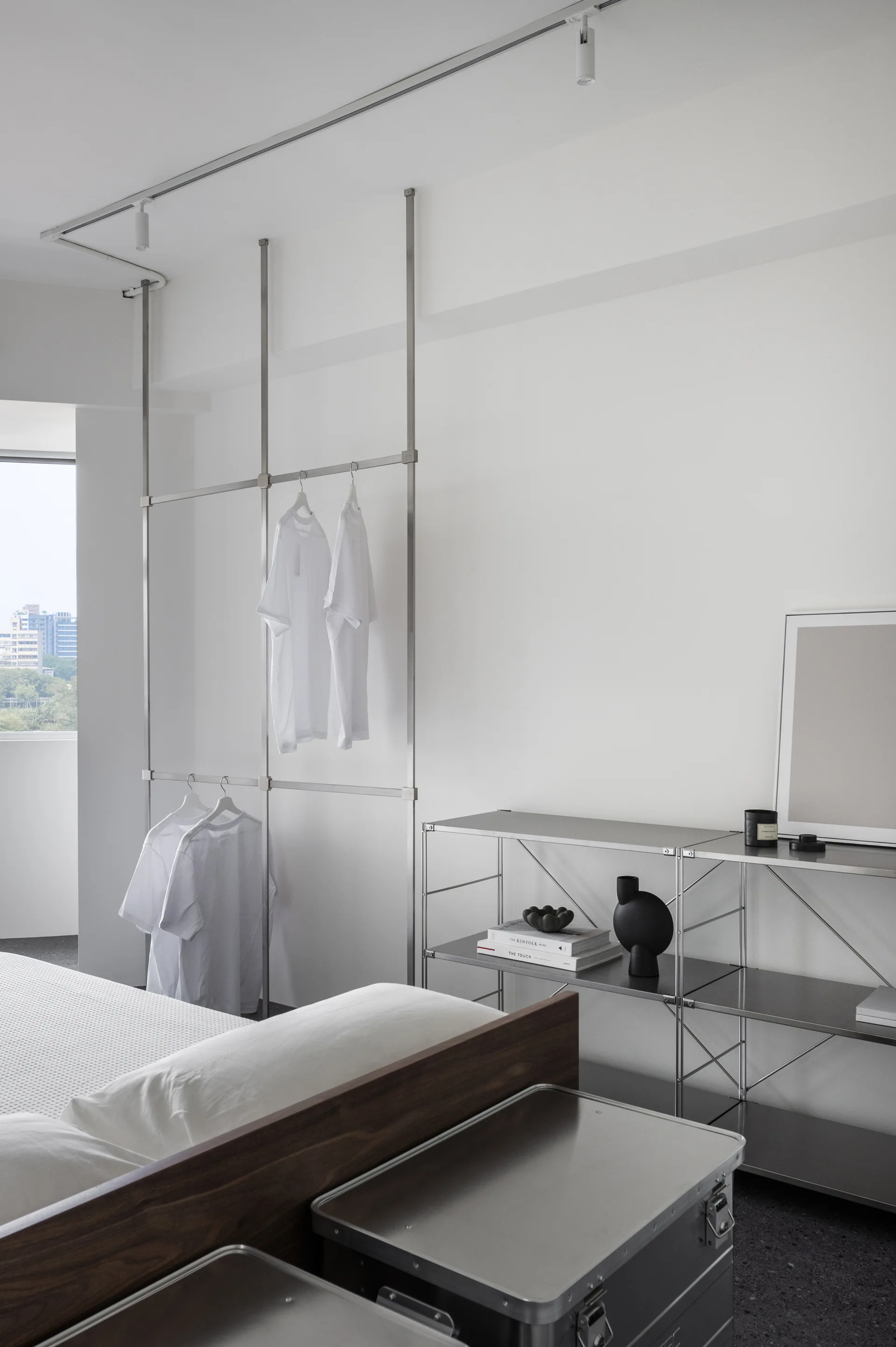 A minimalist bedroom features a metal floor-to-ceiling clothing rack, a modular steel shelving unit, and metallic storage trunks by a wooden bed.