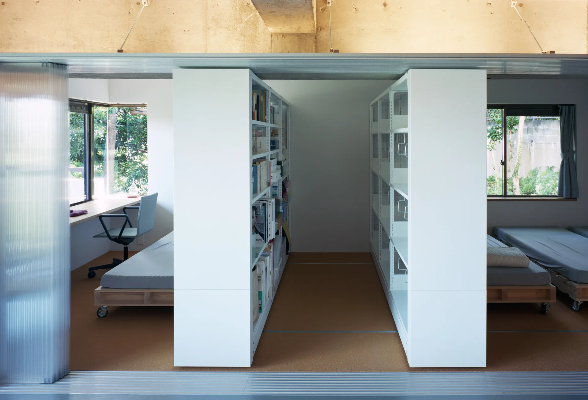 A minimalist shared bedroom and office space featuring tall white bookshelves serving as a partition between platform beds and a wooden desk.