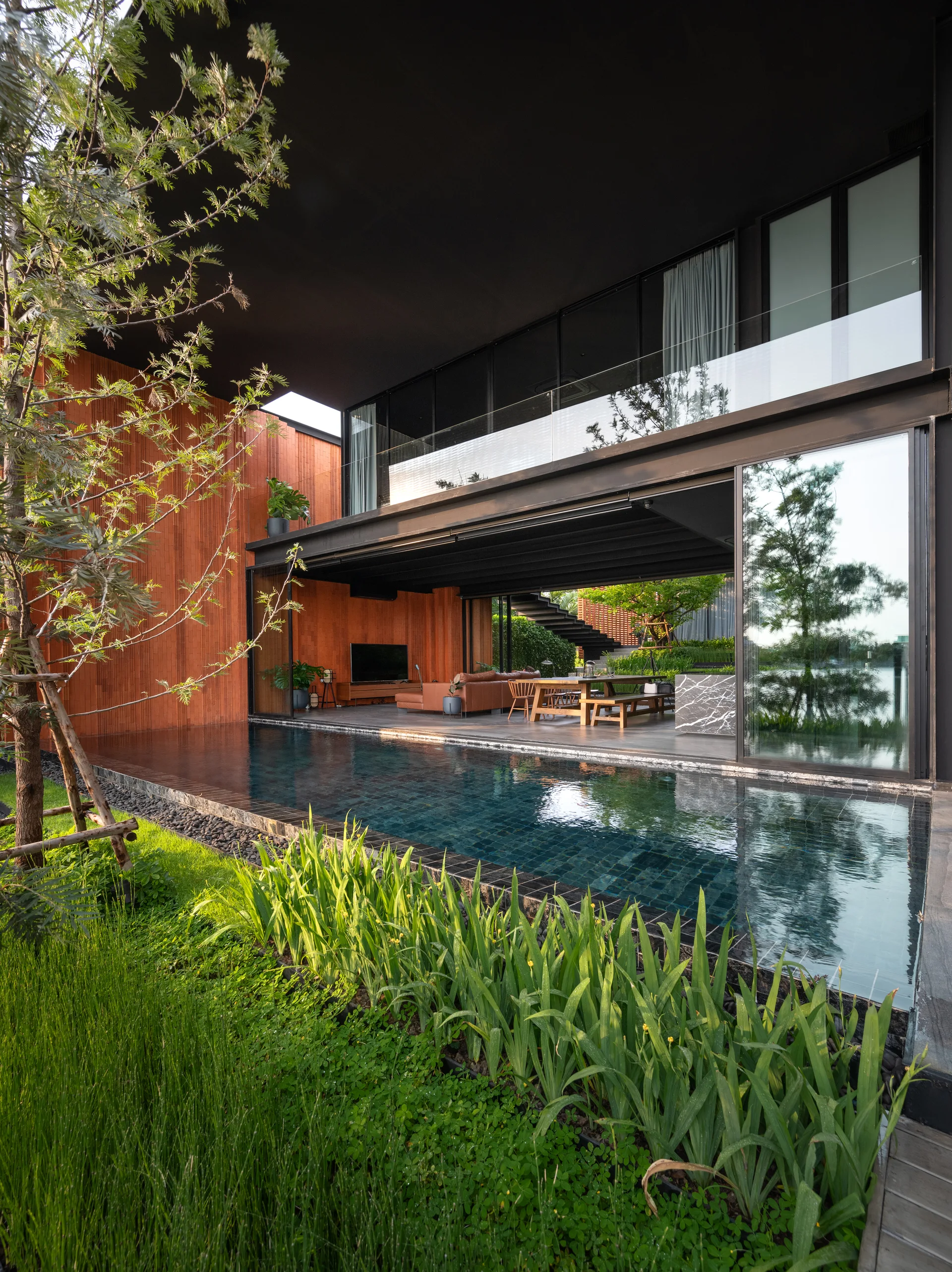 A modern home with a black overhanging ceiling features a living area with sliding glass doors opening onto a swimming pool and grassy garden.