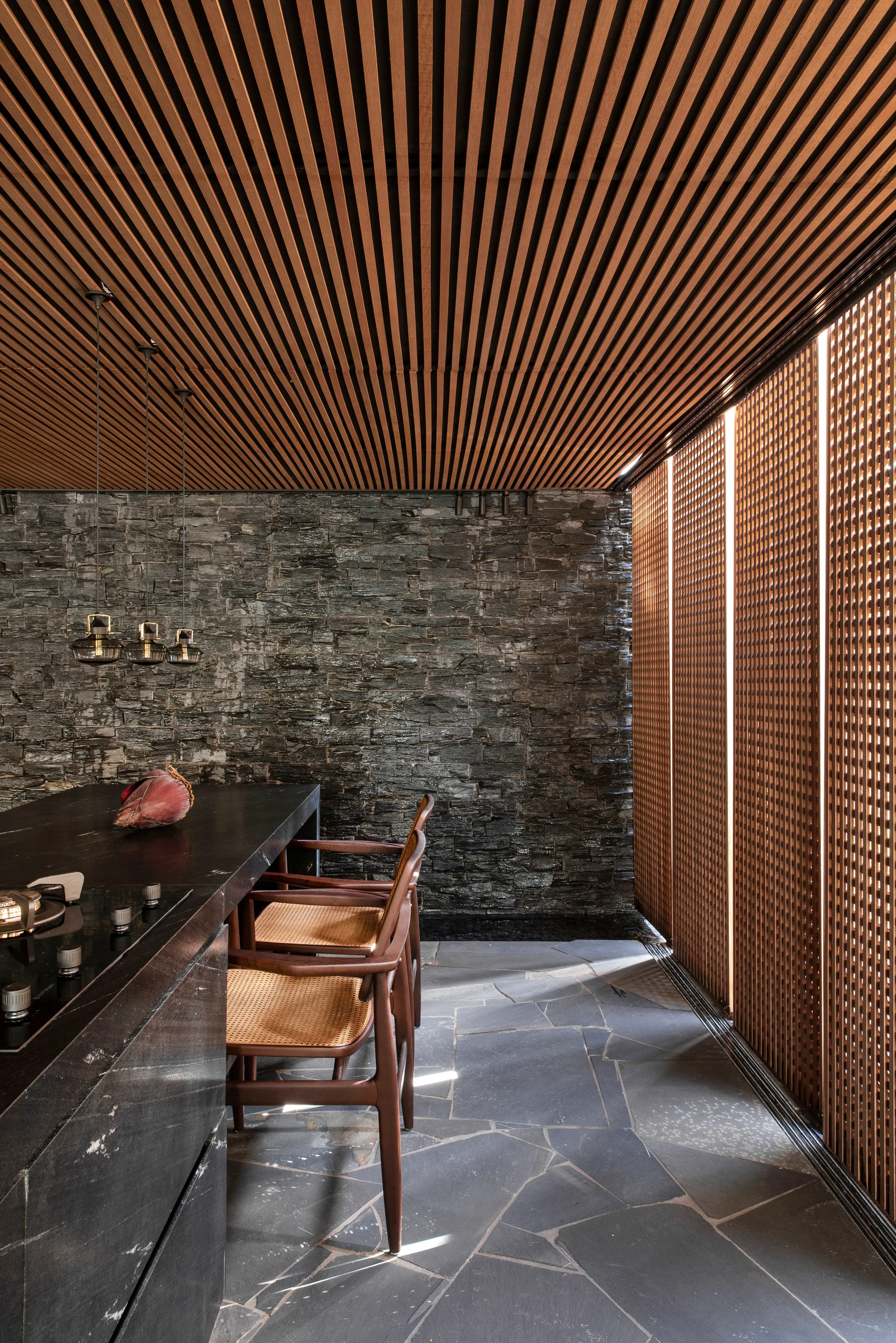 A kitchen with a dark stone island and wooden stools under a slatted ceiling, set against a dark textured stone wall and wood lattice screens.