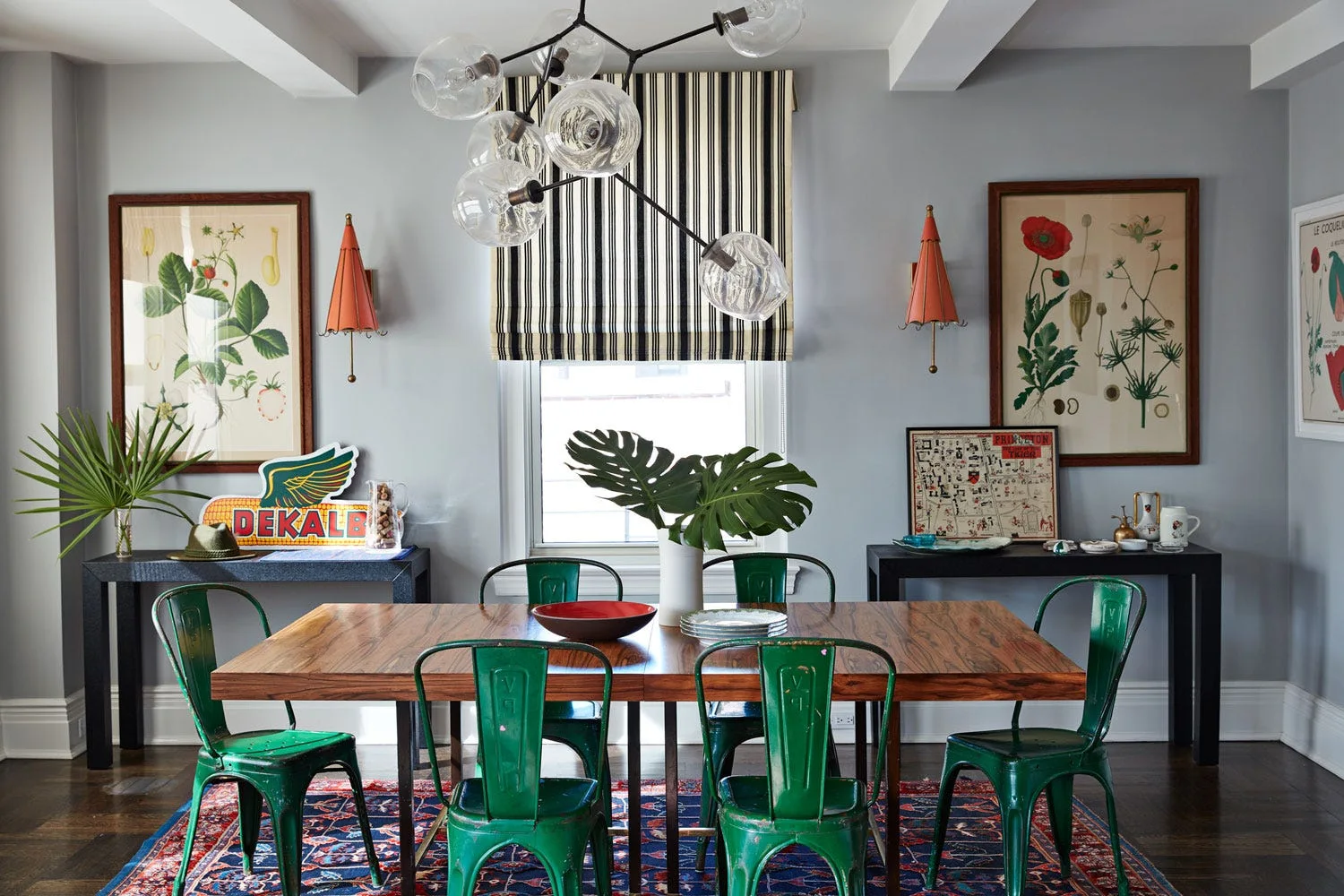 An eclectic dining room features a wooden table, green metal chairs, a striped window shade, and botanical wall art.