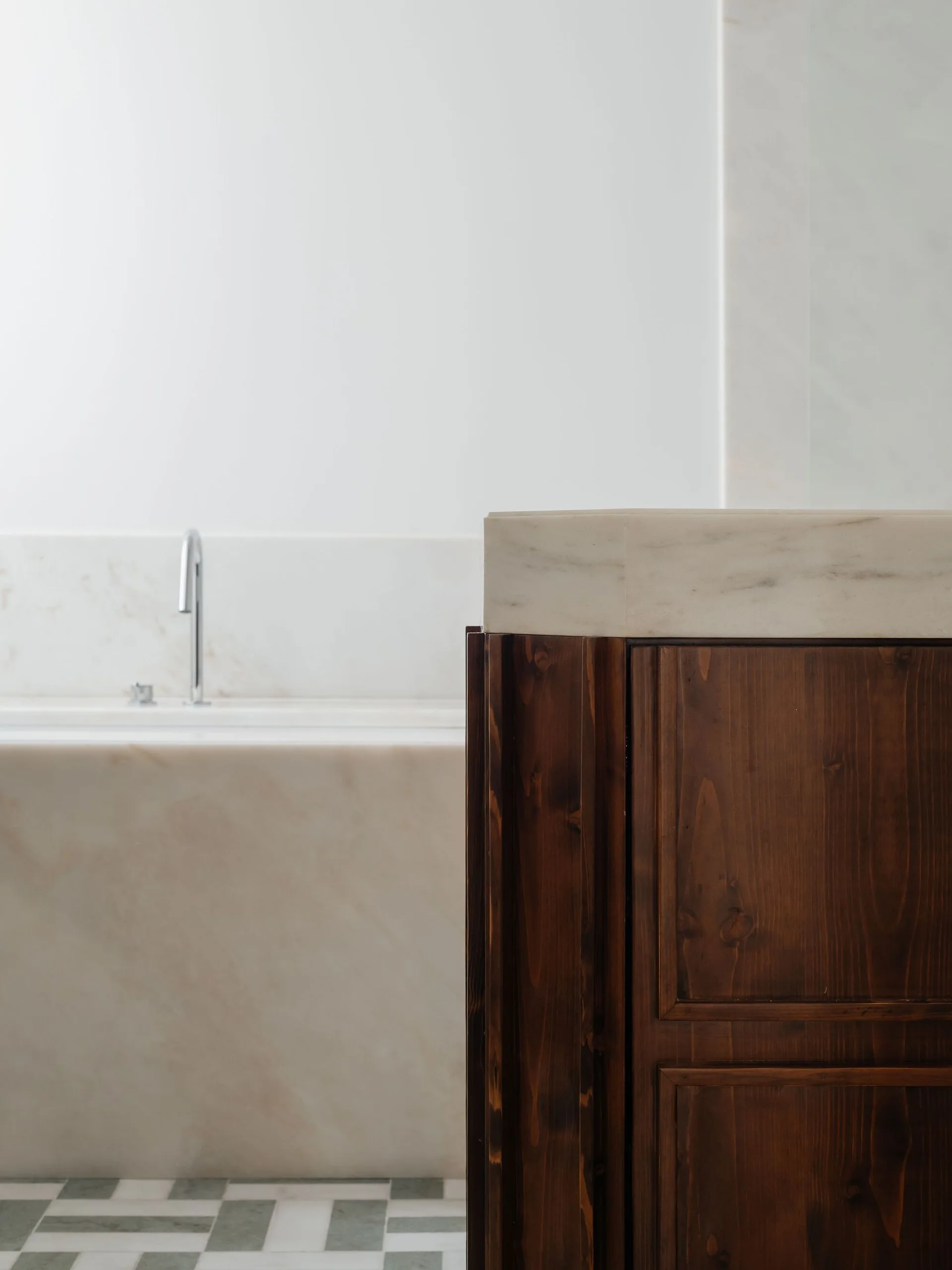 A close-up of a bathroom featuring a dark wood cabinet with a marble top next to a marble-clad bathtub and patterned floor tiles.