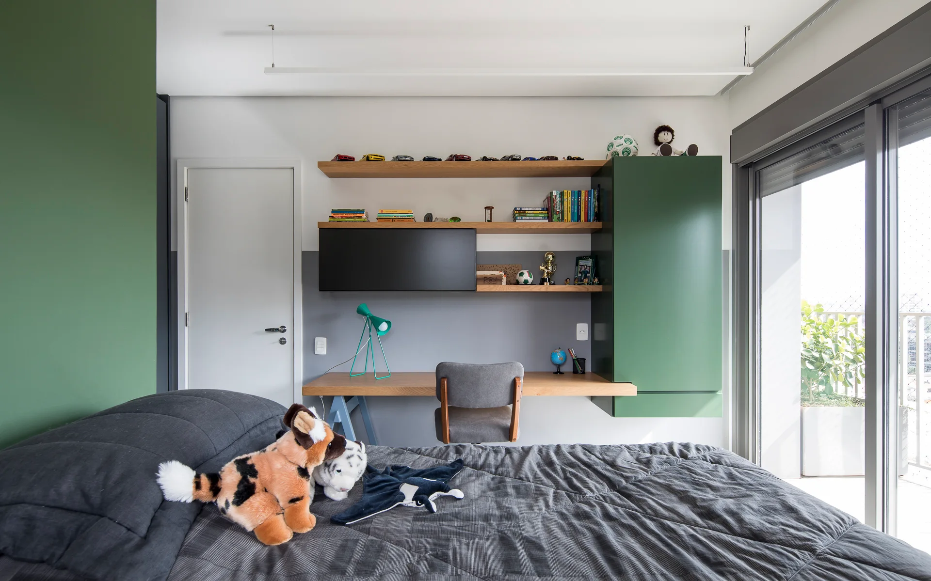 A bedroom features a grey quilted bed in the foreground and a built-in wooden desk with green cabinets and floating shelves on the back wall.
