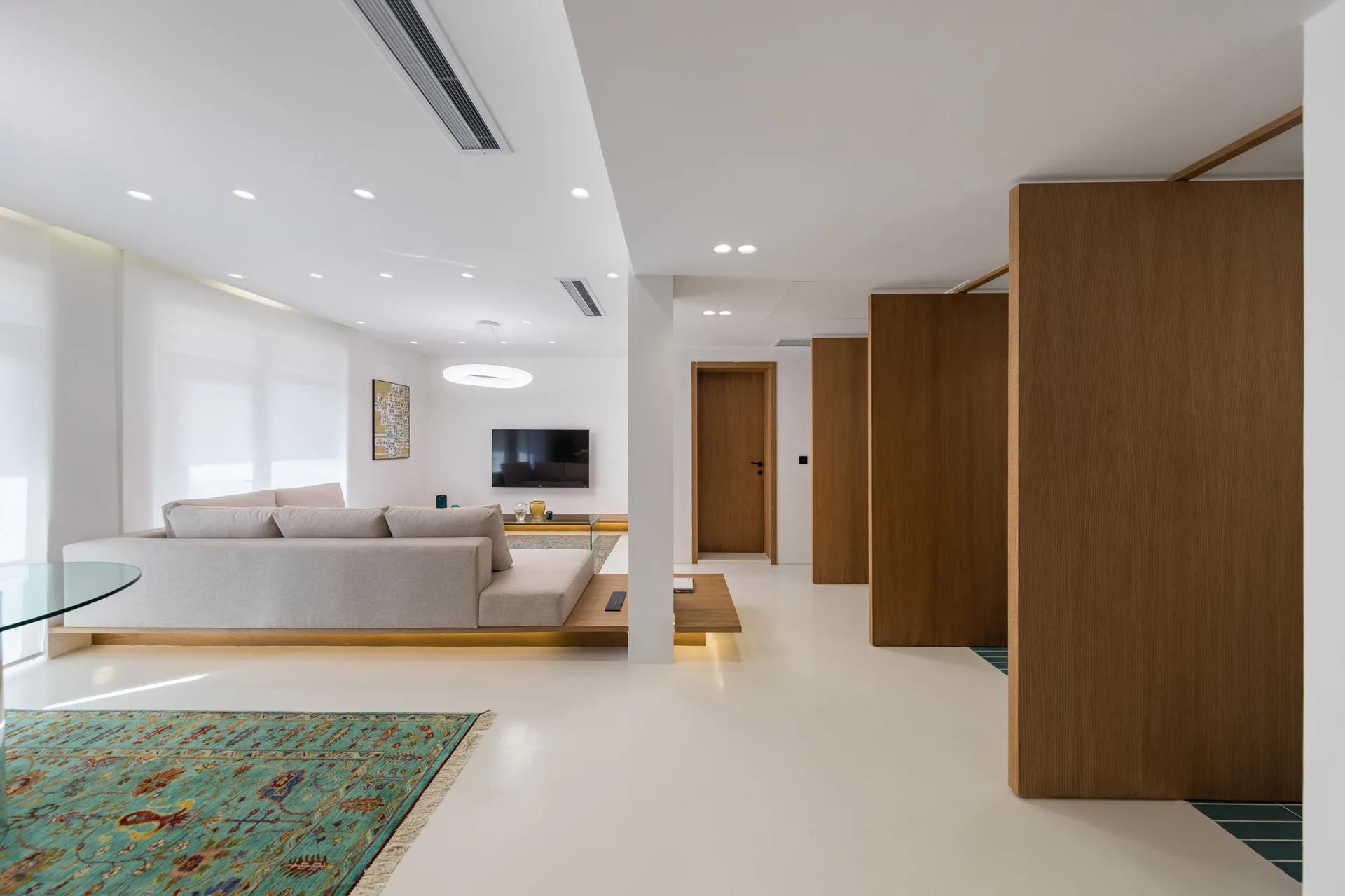 A minimalist living room features a beige modular sofa on a low wooden platform, vertical wood room dividers, and a patterned teal area rug.