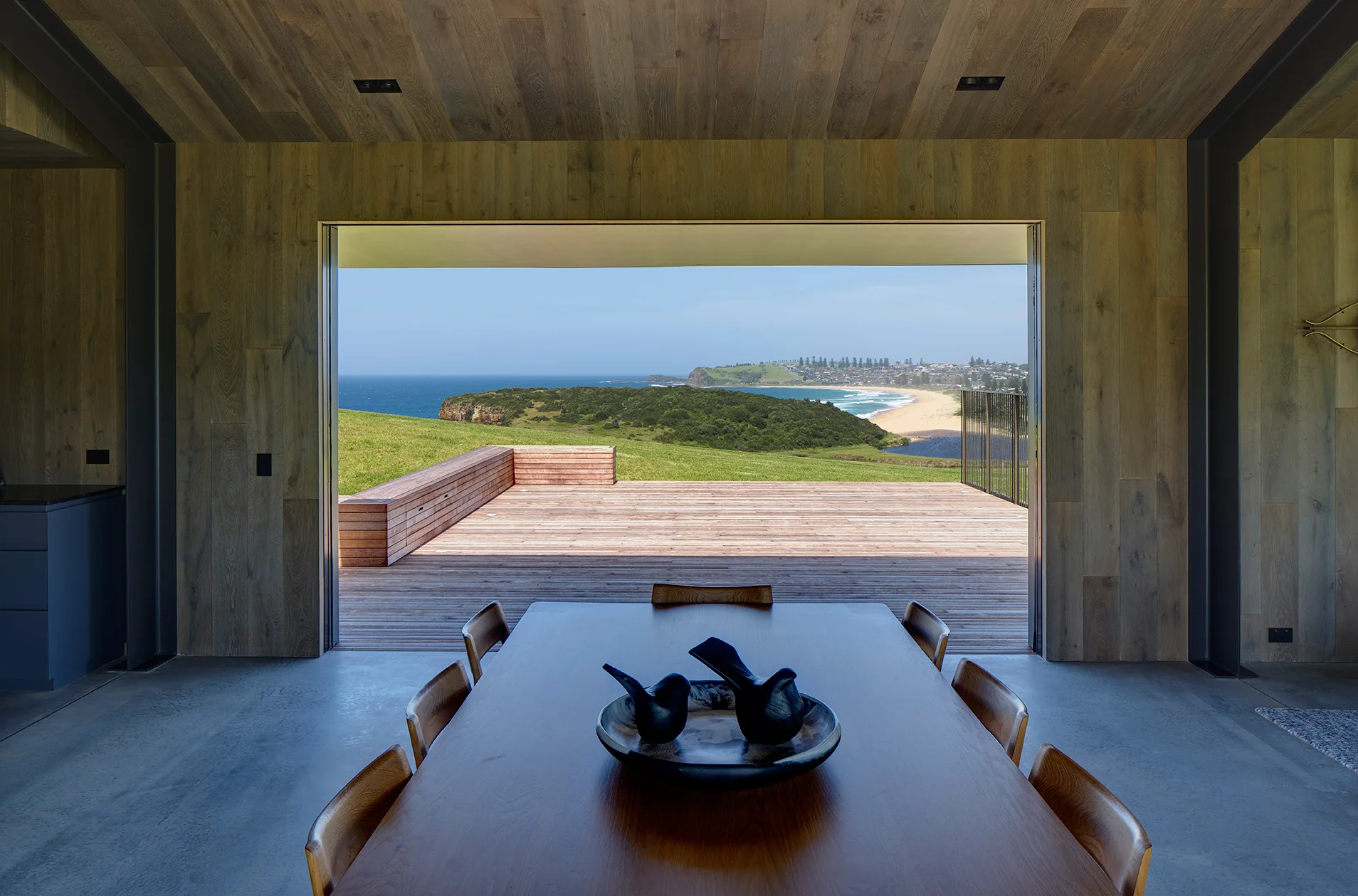 A modern dining room with a long wood table and concrete floor opens onto a spacious wood deck looking out at a grassy coast and ocean.