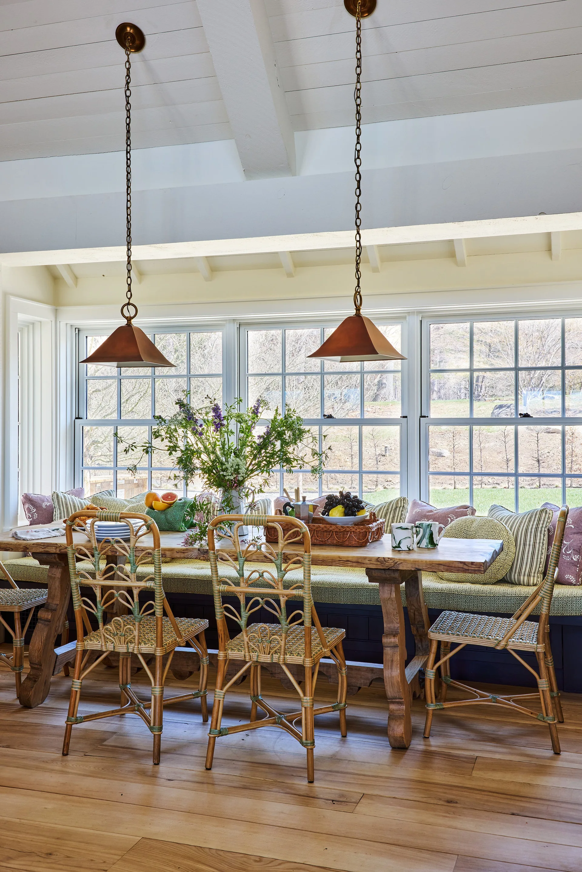 A sunlit dining room features a long wooden table, woven rattan chairs, and a built-in banquette window seat beneath multi-pane windows.