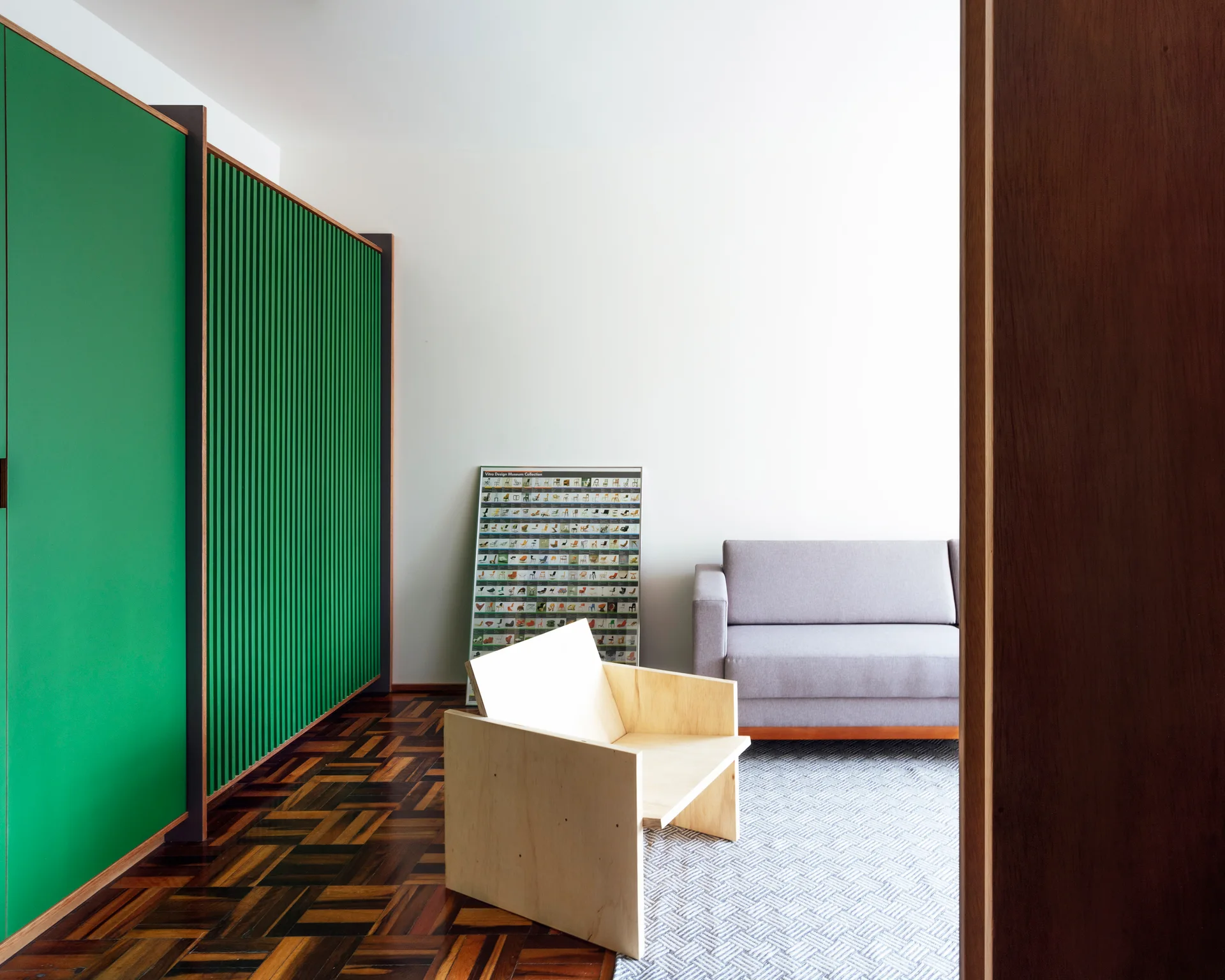 A modern living room features a grey sofa, a geometric light wood armchair, dark parquet flooring, and a large green slatted wall panel.