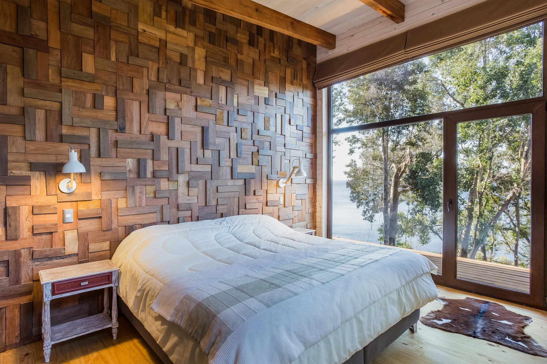 A rustic bedroom features a bed set against a textured wood-block accent wall, flanked by white sconces, next to a large window overlooking trees.
