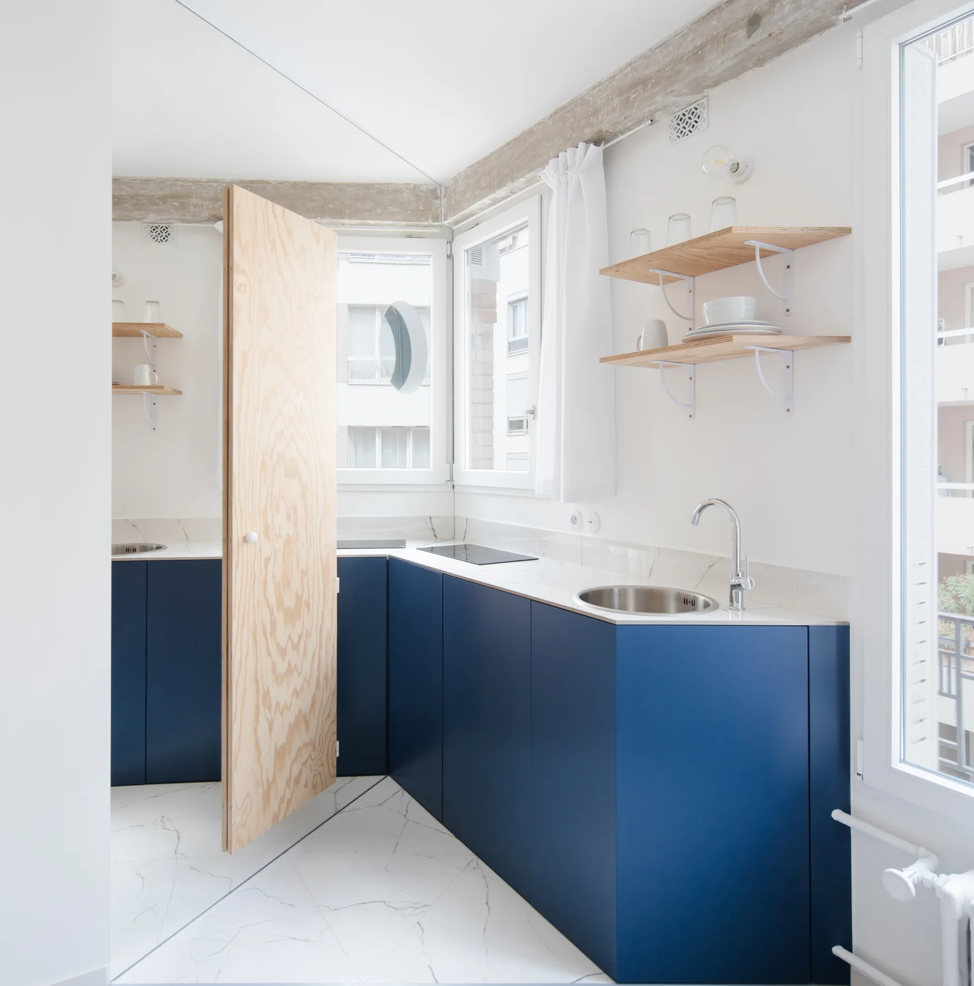 A modern kitchen featuring matte navy blue cabinets, white veined marble flooring, open wood shelves, and an exposed concrete ceiling beam.