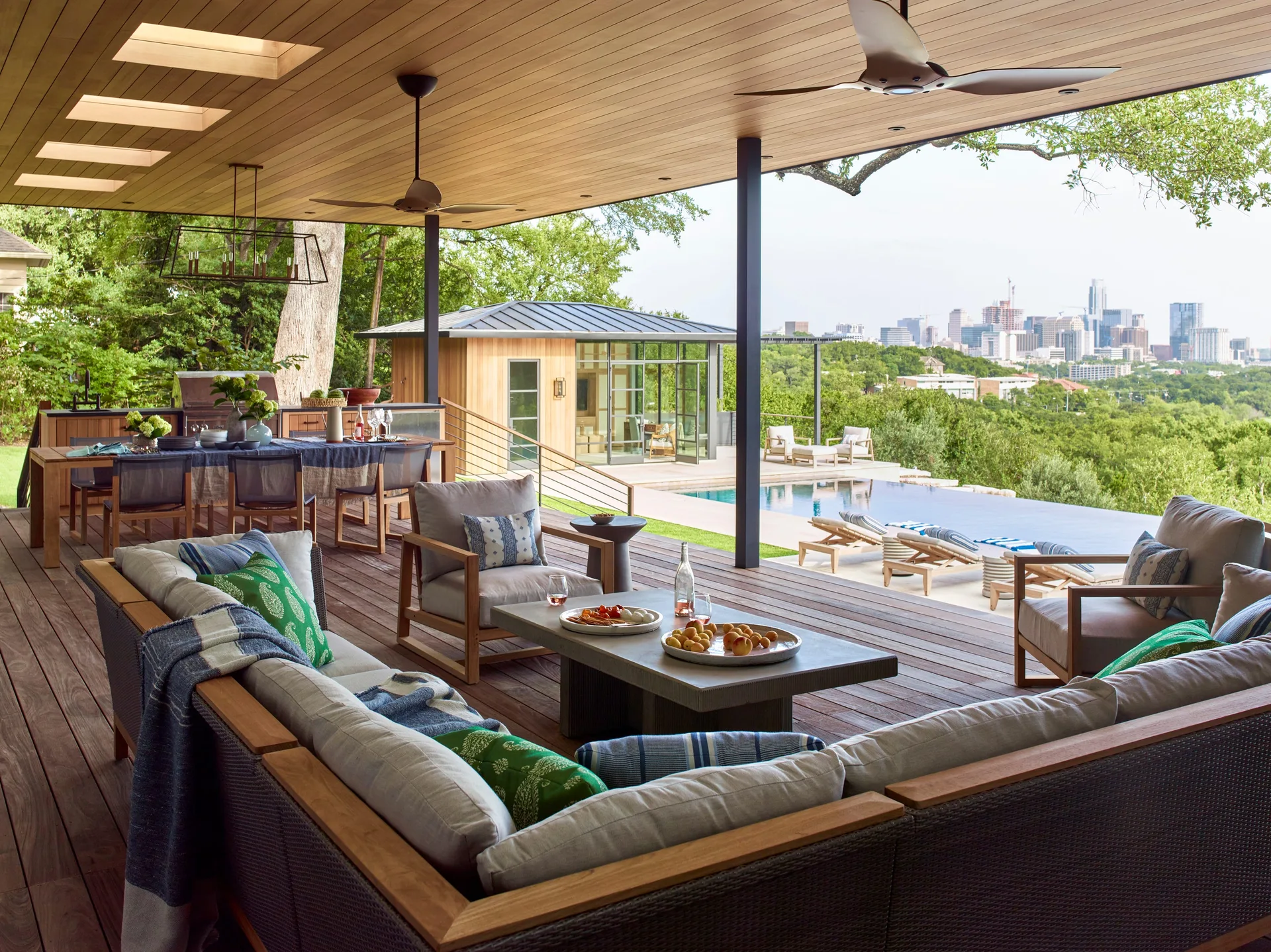 A spacious covered wood patio featuring a grey sectional sofa, an outdoor dining table, and a distant view of an infinity pool and city skyline.