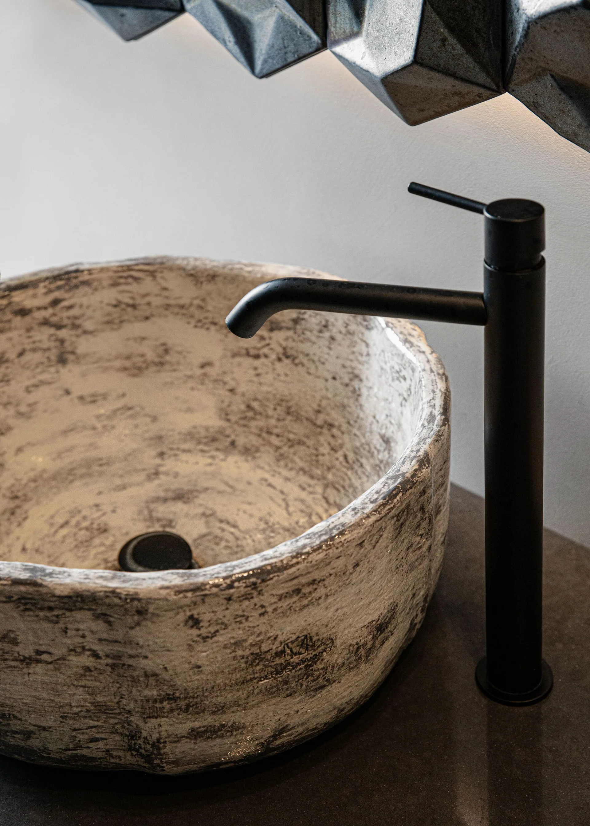 A close-up of a round textured stone vessel sink and a tall matte black faucet on a dark countertop.