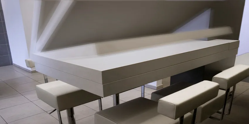 A long minimalist white bar counter is paired with cushioned off-white bar stools featuring shiny chrome bases on a tiled floor.