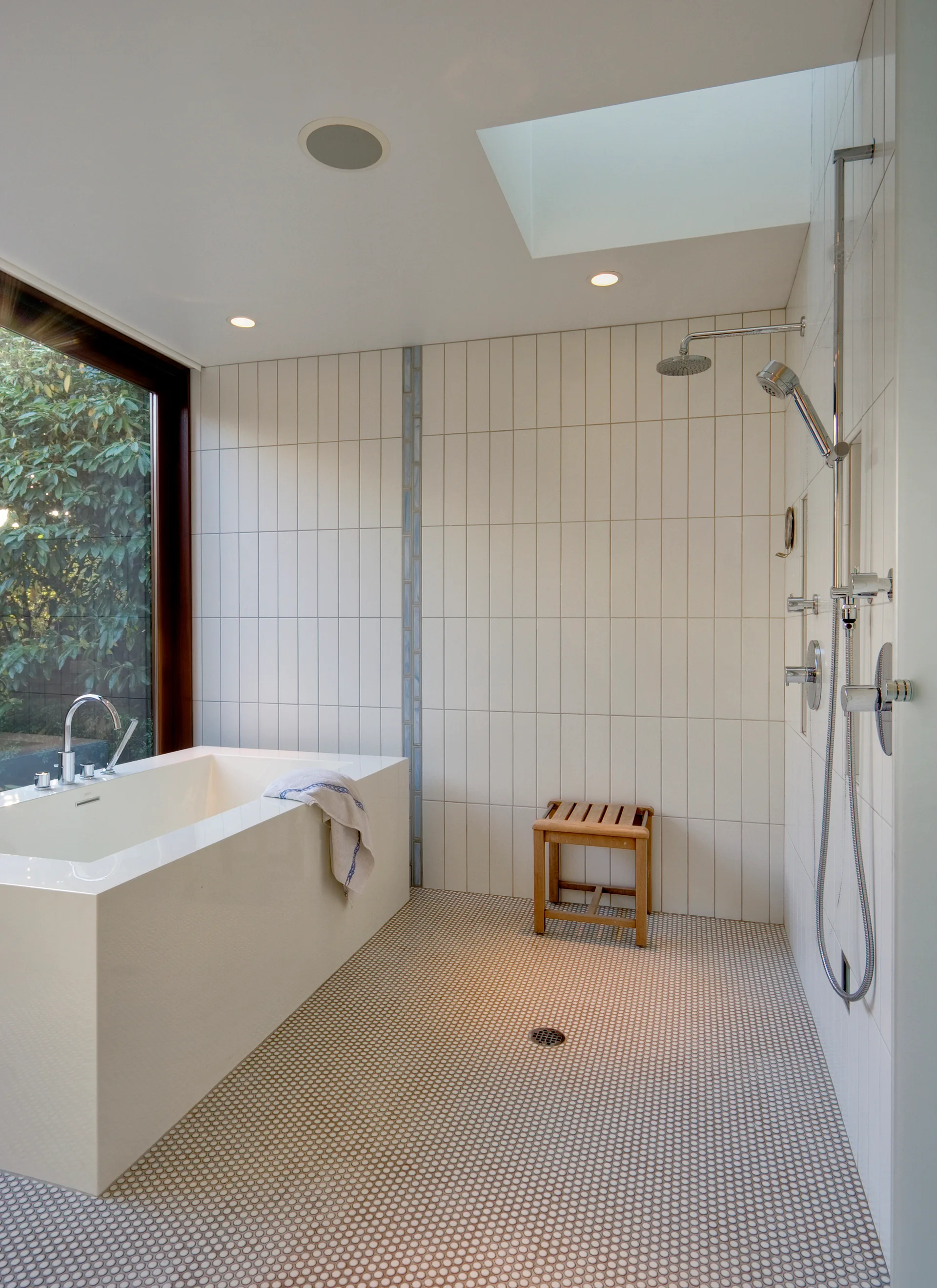 A modern wet-room bathroom featuring a white rectangular bathtub, vertical wall tiles, a penny round mosaic floor, and a wooden stool.