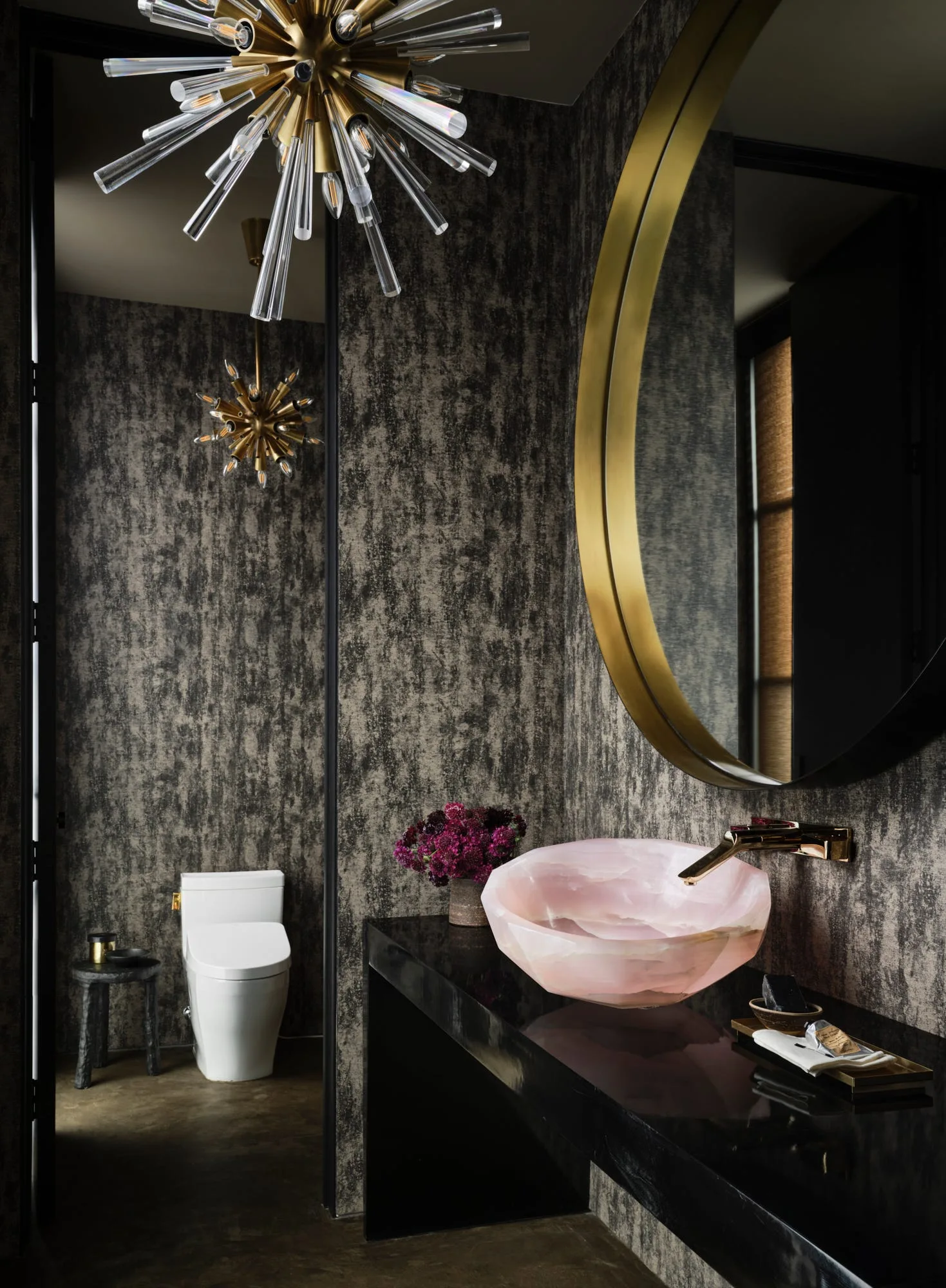A moody powder room with mottled dark wallpaper, a pink stone vessel sink, a glossy black vanity, and a spiky gold chandelier.