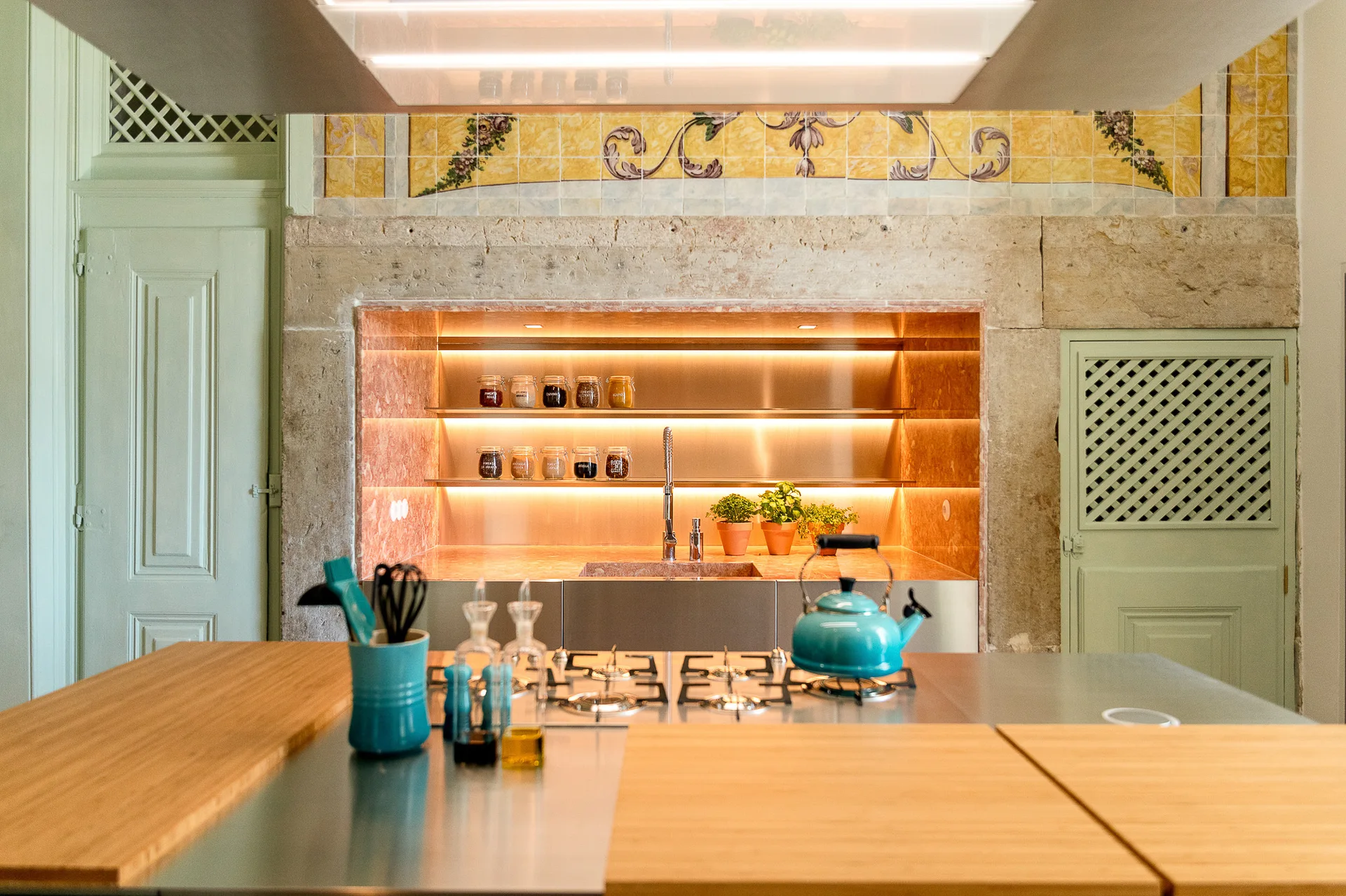 A modern metallic kitchen island with a gas stovetop stands before a stone-framed wall niche with lighted shelves and yellow ornate wall tiles.