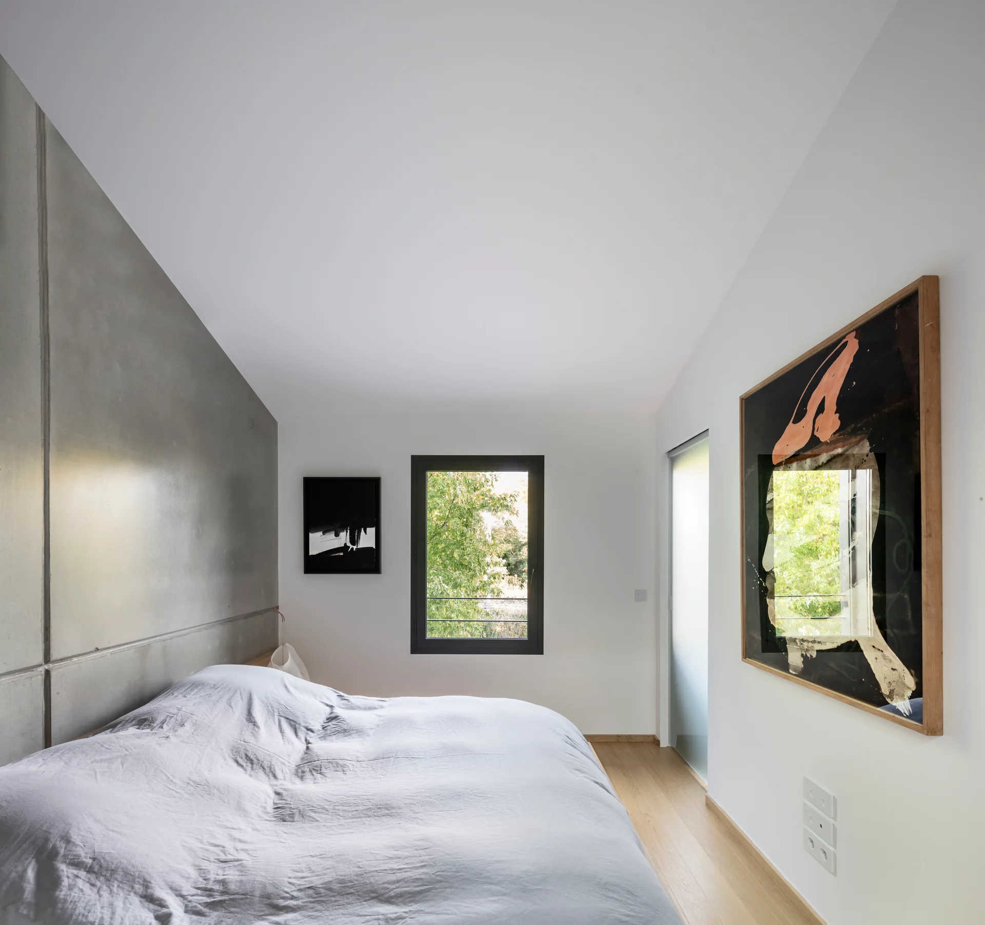 A minimalist bedroom featuring a grey concrete accent wall behind a bed with grey linens, wood flooring, and framed artwork.