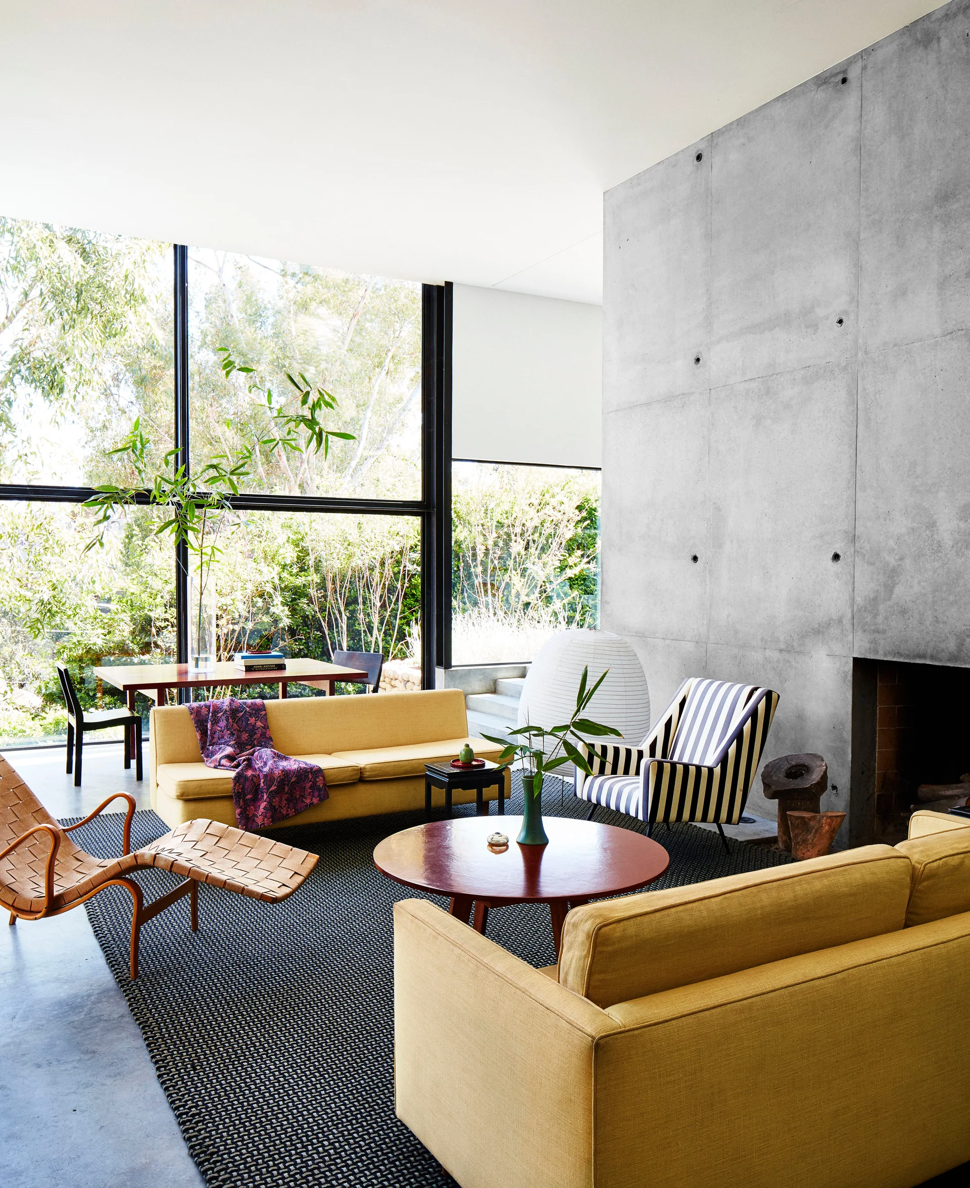 A modern living room features two yellow sofas, a striped armchair, and a woven lounge chair facing an industrial concrete fireplace wall.
