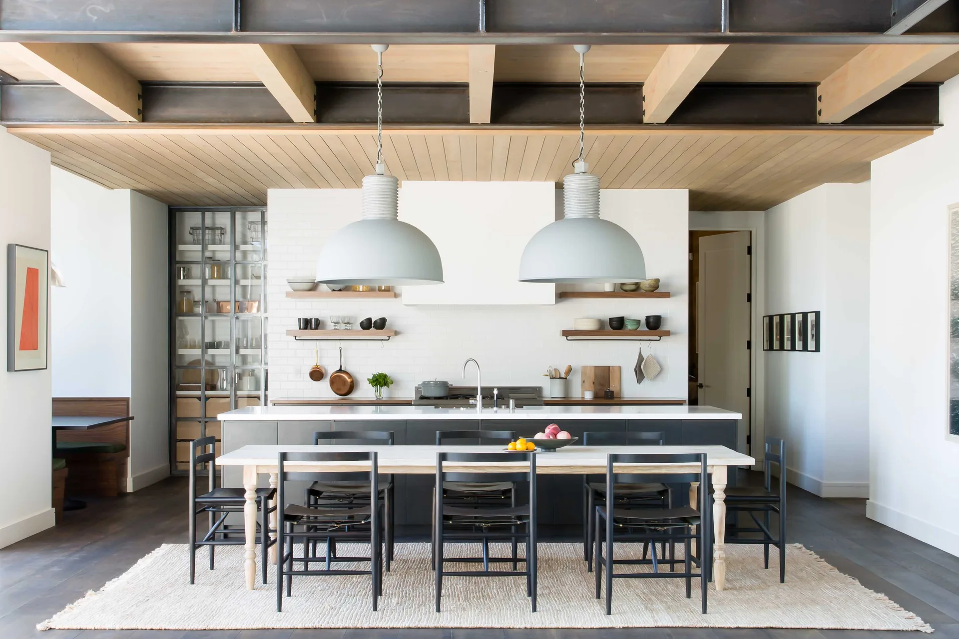 An industrial kitchen features a dark island, a wooden dining table on a textured rug, and grey pendant lights under an exposed beam ceiling.