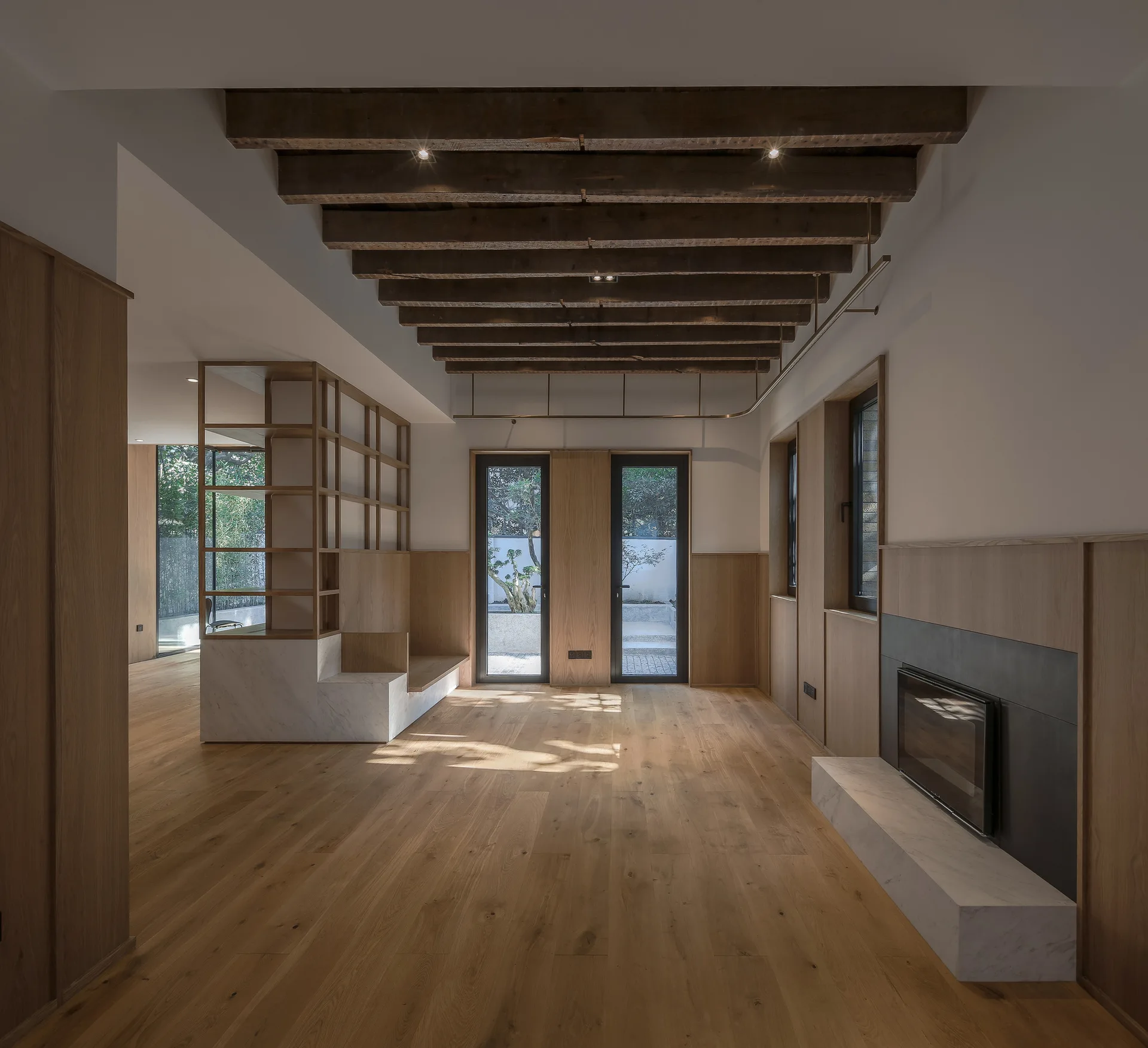 An empty minimalist living room featuring exposed wood ceiling beams, wood wall paneling, and a fireplace with a low marble hearth.