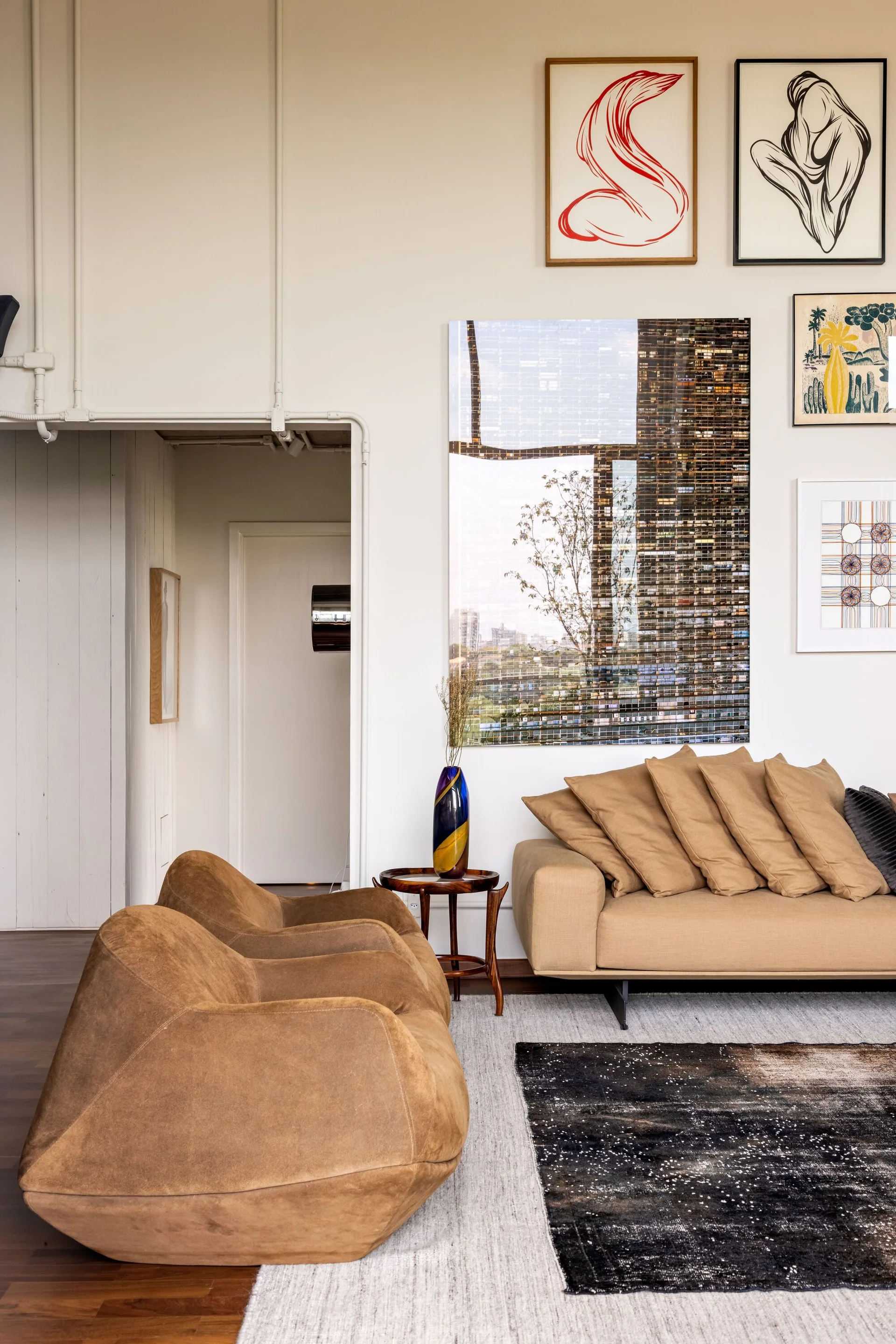 A contemporary living room featuring tan suede armchairs, a matching sofa with stacked pillows, a gallery wall of art, and layered area rugs.