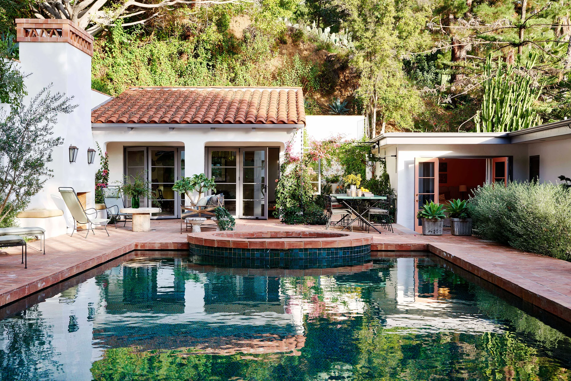 A backyard swimming pool with dark teal water is surrounded by a terracotta tile terrace, white stucco walls, and a tall outdoor fireplace.