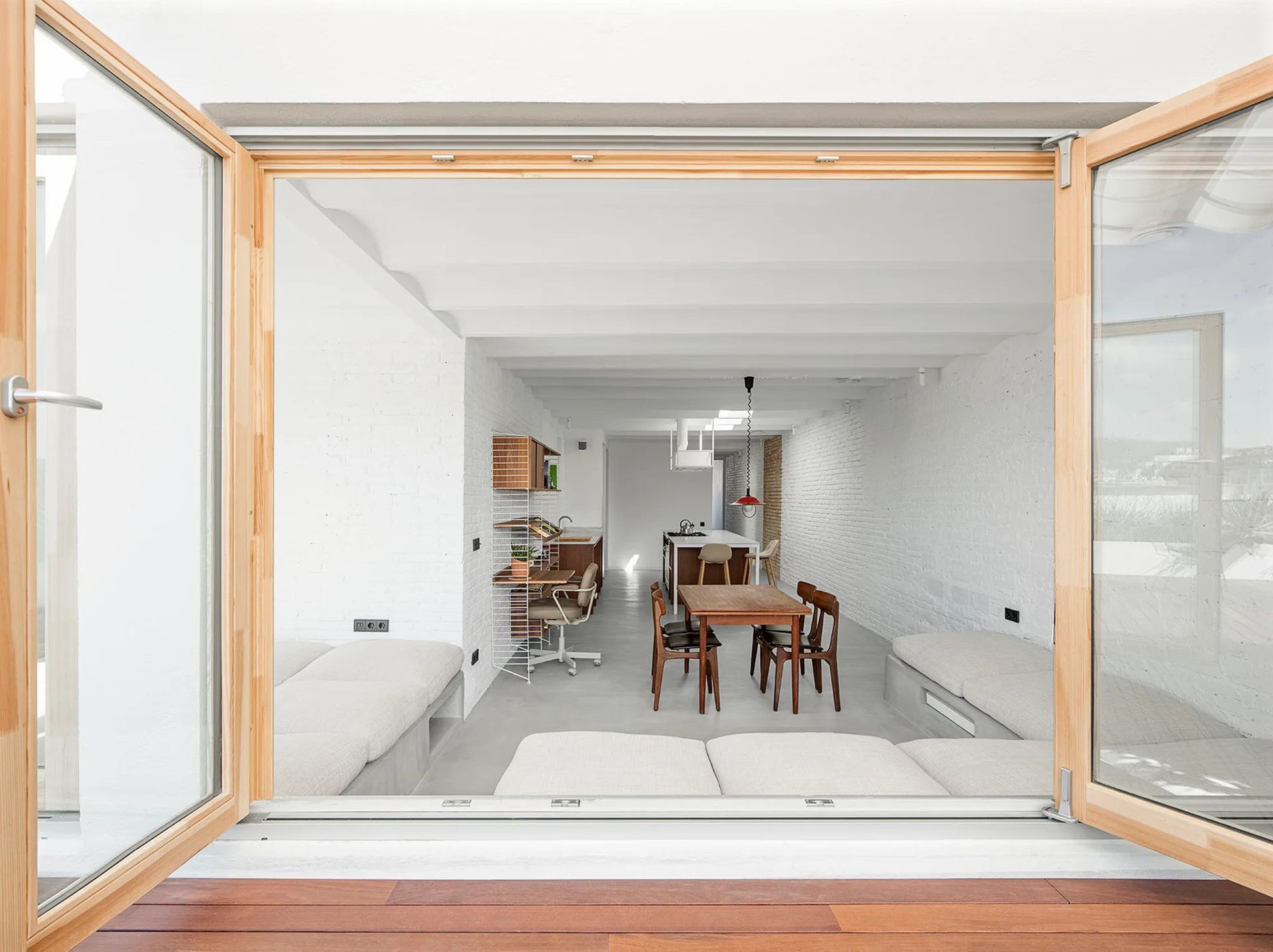 A view through open wooden double doors into a minimalist living space with white painted brick walls, concrete floors, and a dining table.