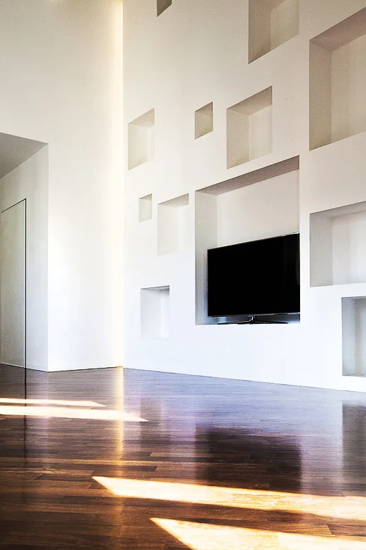 A large white minimalist wall features multiple recessed square niches and an integrated television, set above glossy dark hardwood floors.