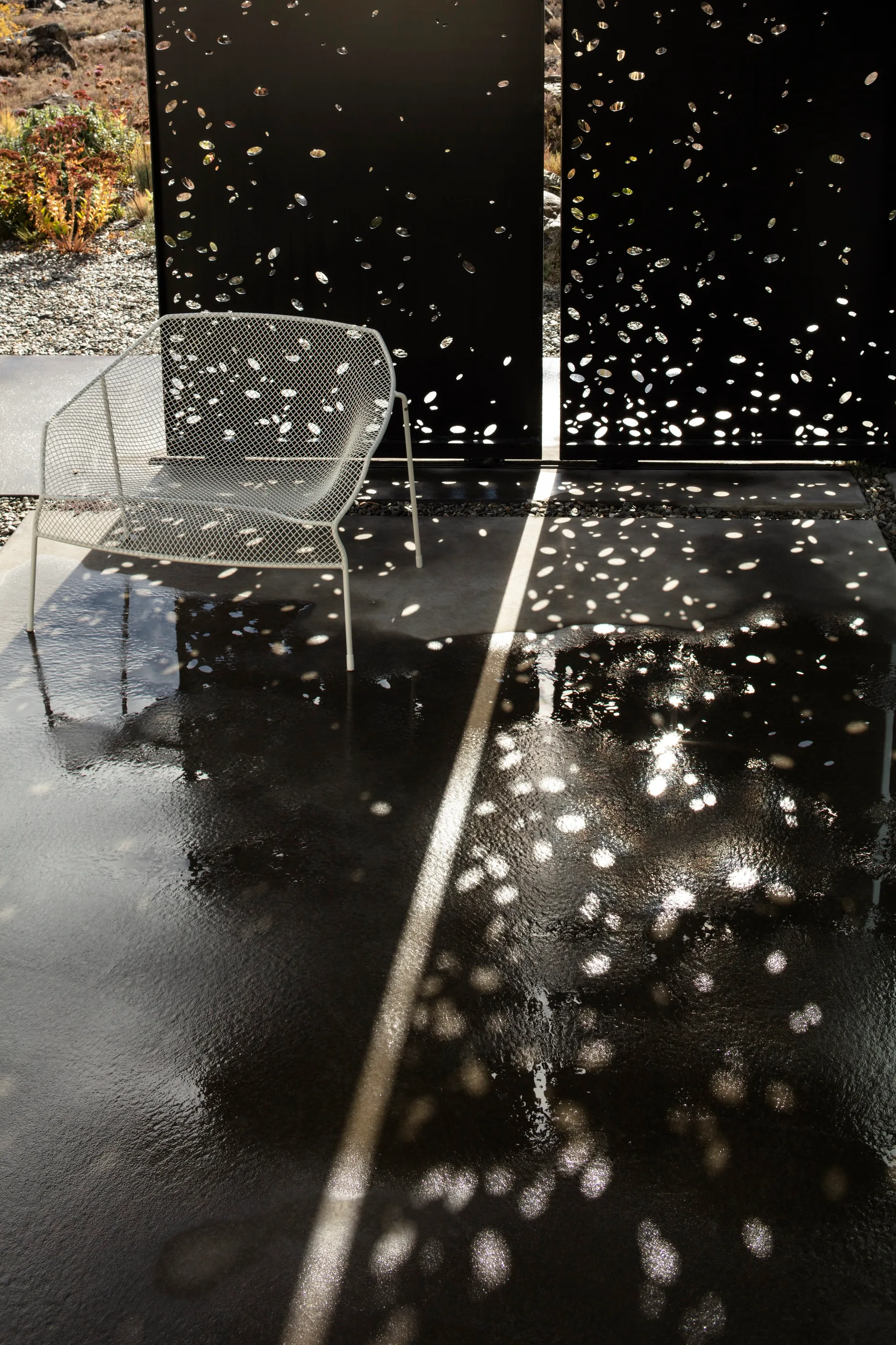 A white metal mesh lounge chair rests on a wet concrete patio in front of a black perforated metal privacy screen casting dappled shadow patterns.