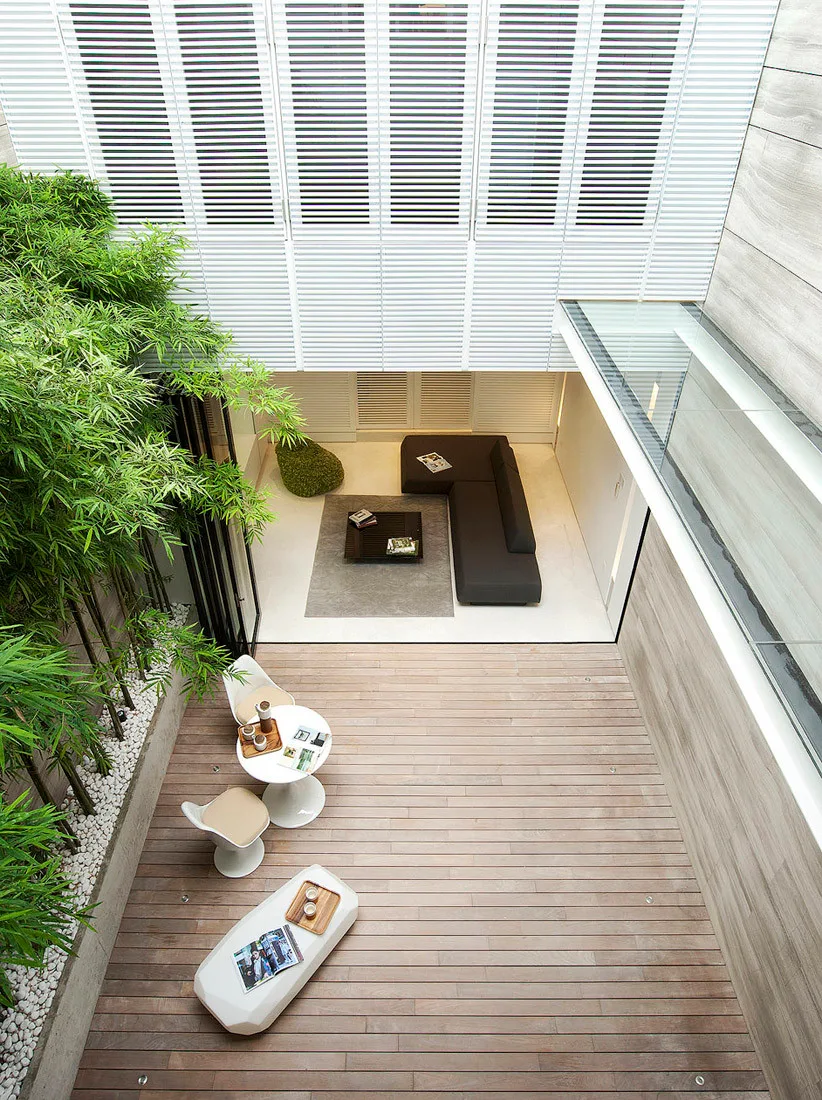 A high-angle view of an outdoor courtyard with light wood decking, tall bamboo plants, and a transition to an indoor living room with a dark sectional.