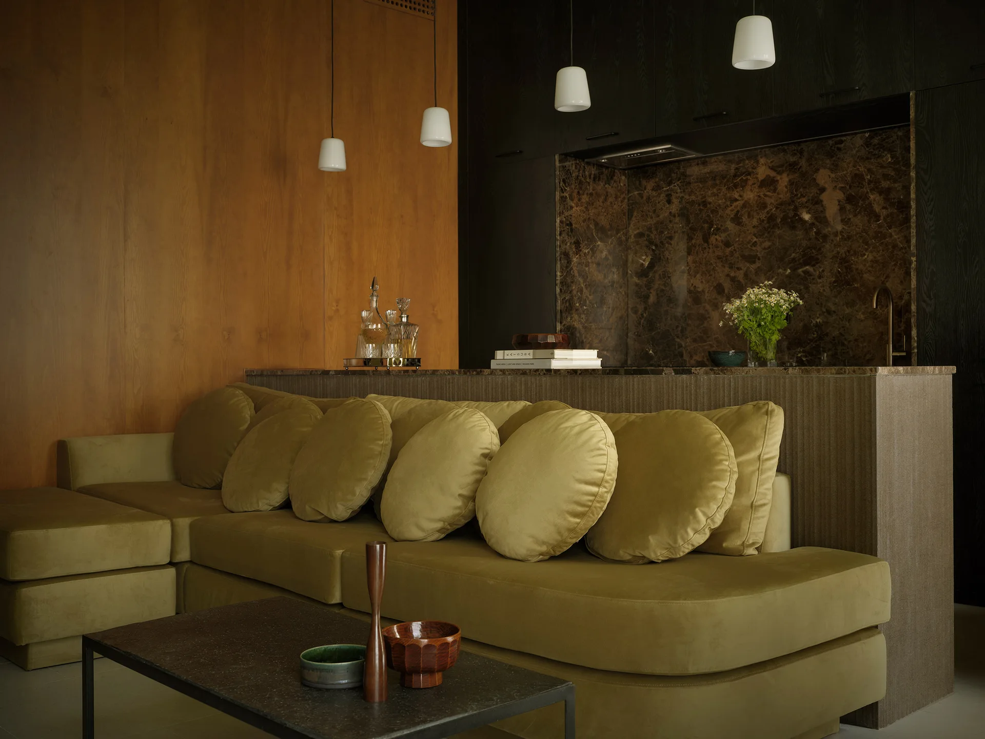An olive green velvet sectional sofa with round pillows sits in front of a kitchen island with a dark stone backsplash and wood wall paneling.