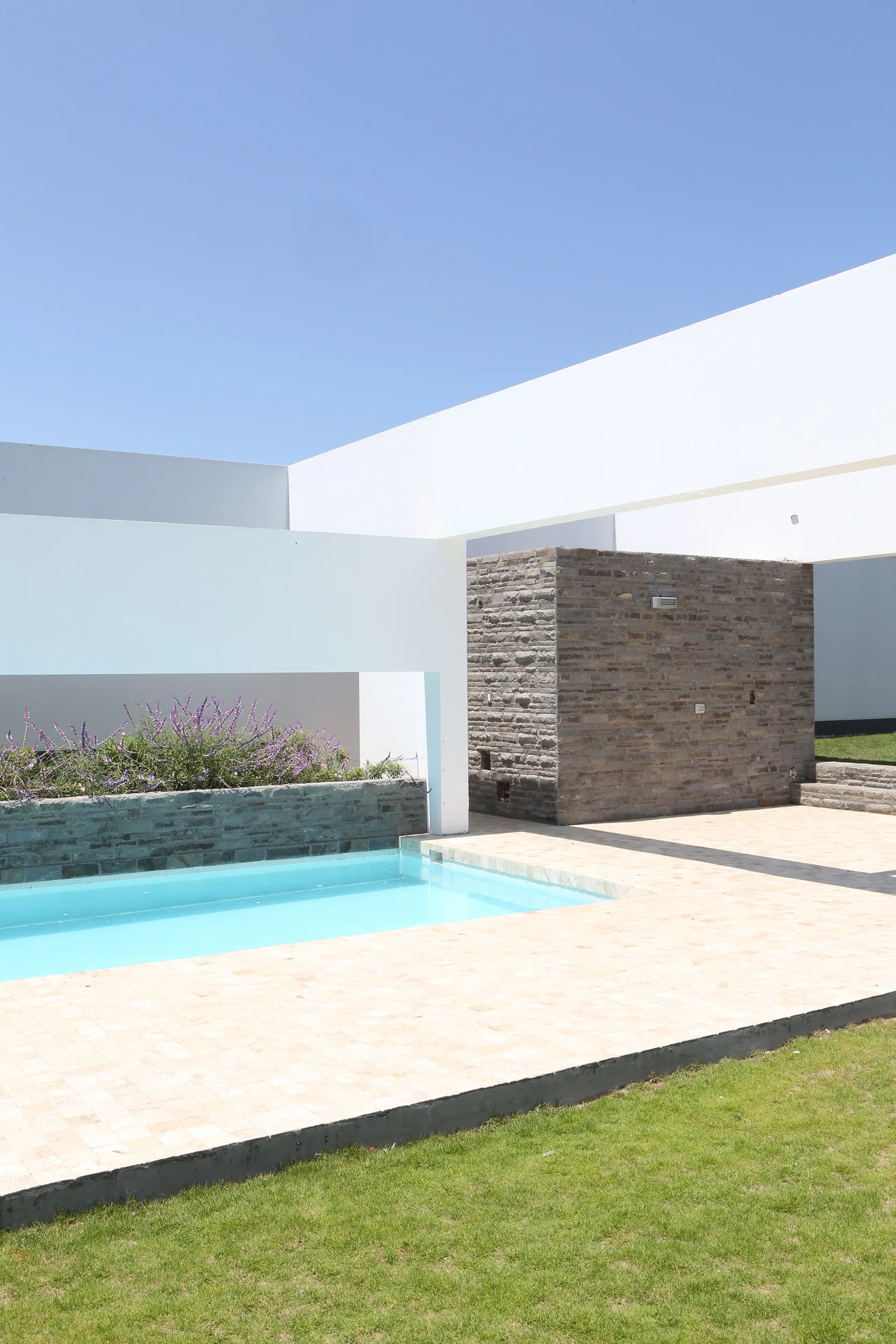 A rectangular light blue swimming pool surrounded by beige tile decking, a textured grey stone wall, and white geometric concrete beams.