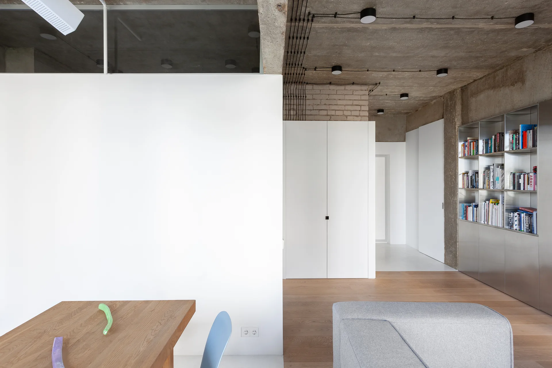 A modern apartment interior features an exposed concrete ceiling with black light fixtures, a white partition wall, wood flooring, and a metal bookcase.