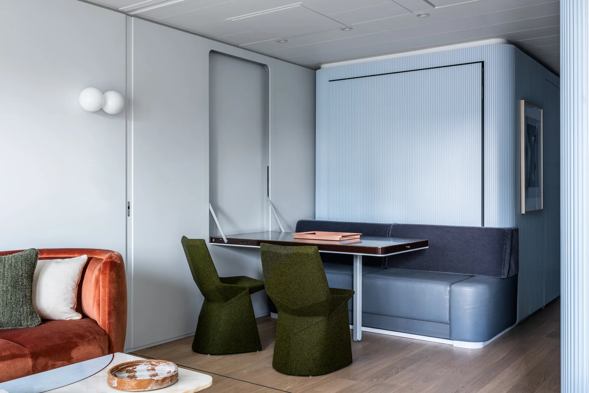 A modern dining area features a fold-down wall table, a blue banquette against fluted panels, green chairs, and a terracotta velvet sofa.
