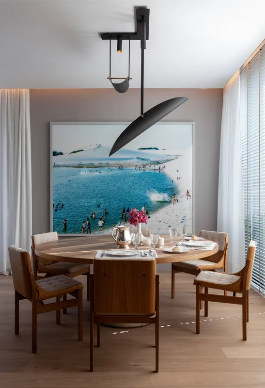 A contemporary dining room with a round wooden table, chairs, a black sculptural pendant light, and a large beach photo on the wall.