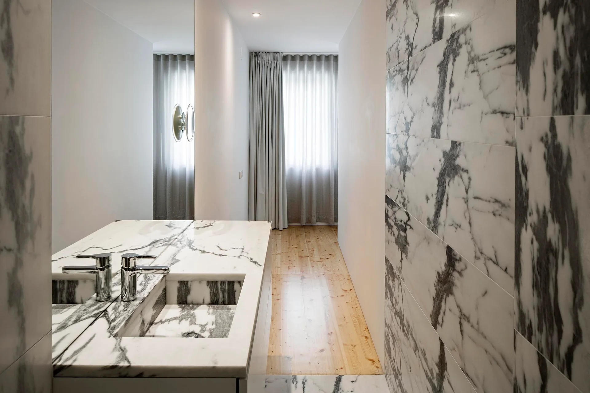 A minimalist bathroom with polished marble wall tiles, an integrated marble vanity sink, a large mirror, and light wood flooring.