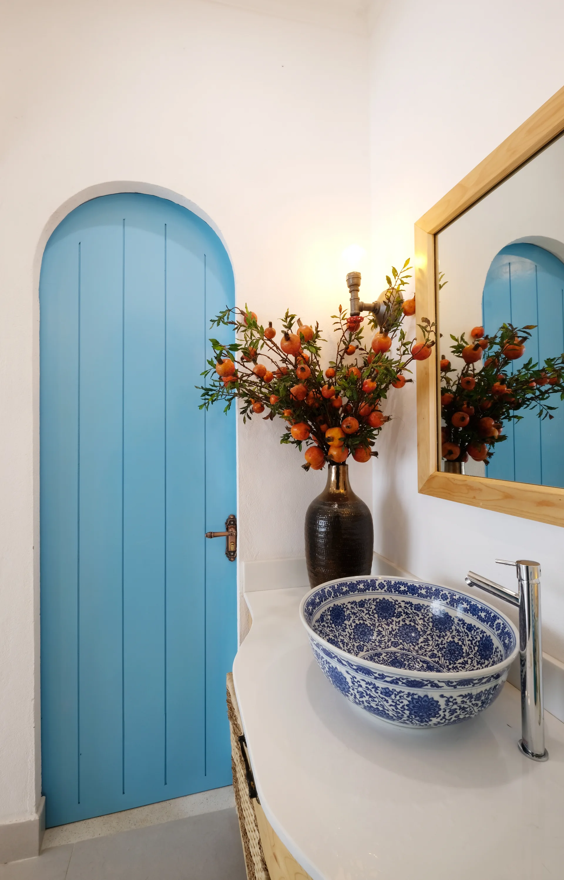 A bathroom vanity featuring a blue patterned ceramic vessel sink, a light wood-framed mirror, and an arched light blue door set in a white wall.
