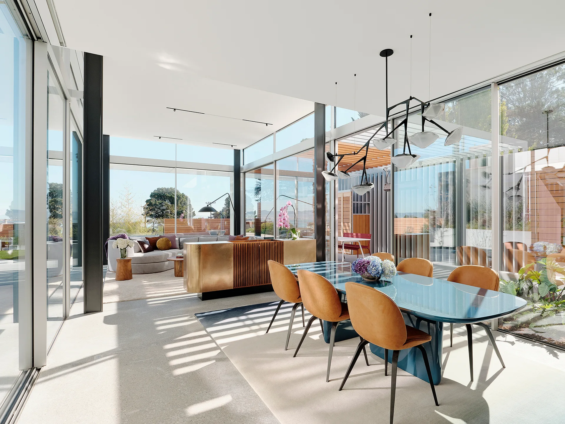 A modern open dining space features a glossy blue table paired with tan leather chairs beneath a geometric black chandelier next to glass walls.