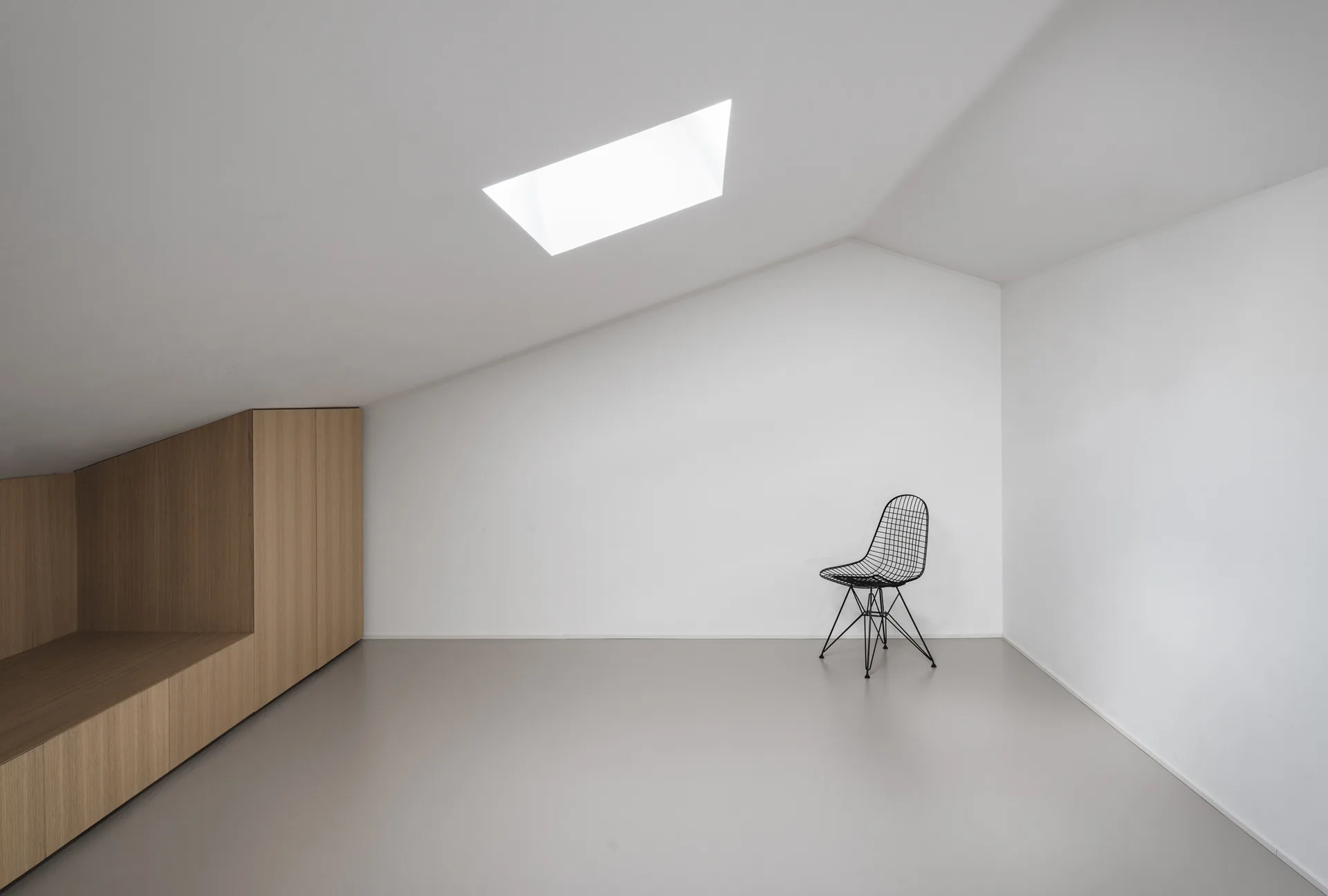 A minimalist attic room features a sloped white ceiling with a skylight, light wood built-in cabinets, a light grey floor, and a single black wire chair.