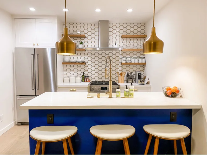 A kitchen features a royal blue island with bar stools, gold dome pendant lights, and a white hexagonal tile backsplash with wood floating shelves.