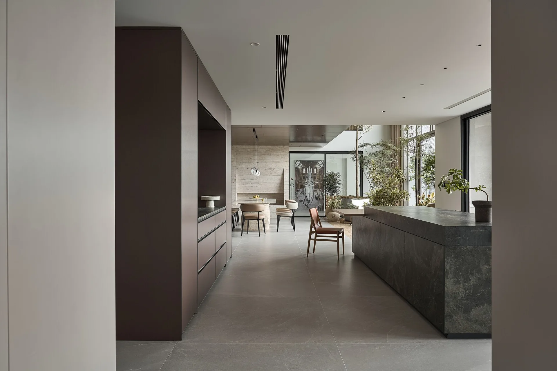 A spacious minimalist kitchen features a large dark stone island, full-height matte brown cabinetry, and grey tile flooring leading to a dining area.