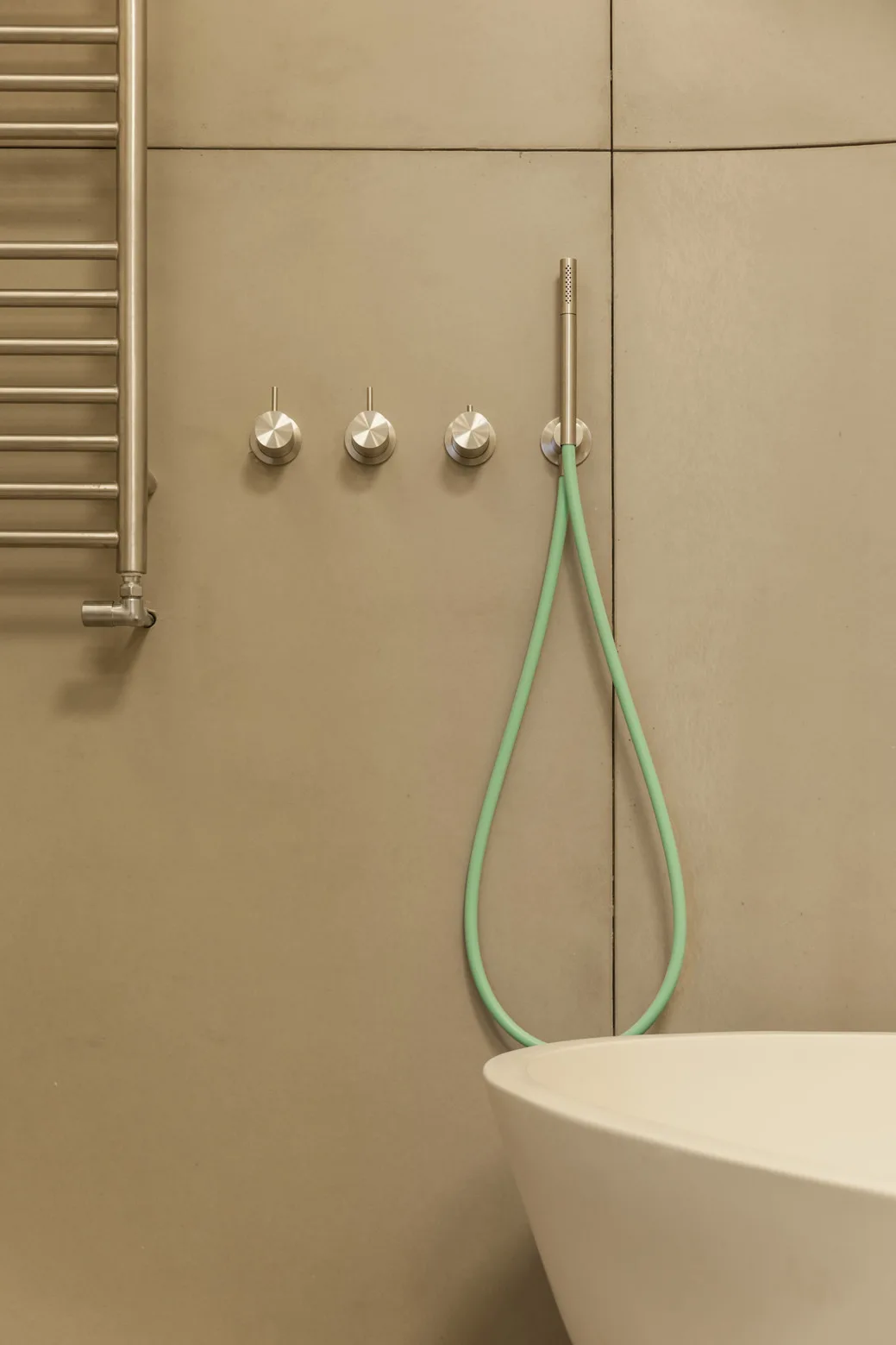 A close-up of a beige paneled bathroom wall featuring a brushed metal towel warmer, three circular controls, and a handheld shower with a light green hose.