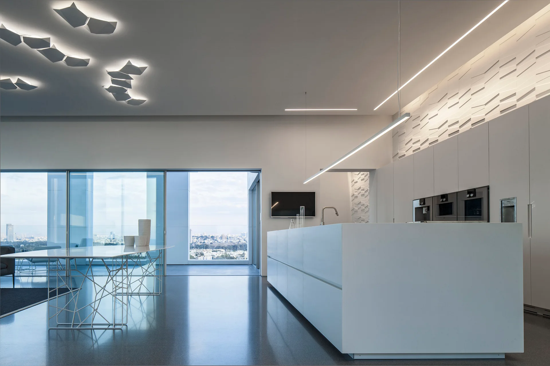 A sleek white kitchen with a large central island, integrated cabinetry, a 3D geometric wall panel, and a dining table with wire legs.