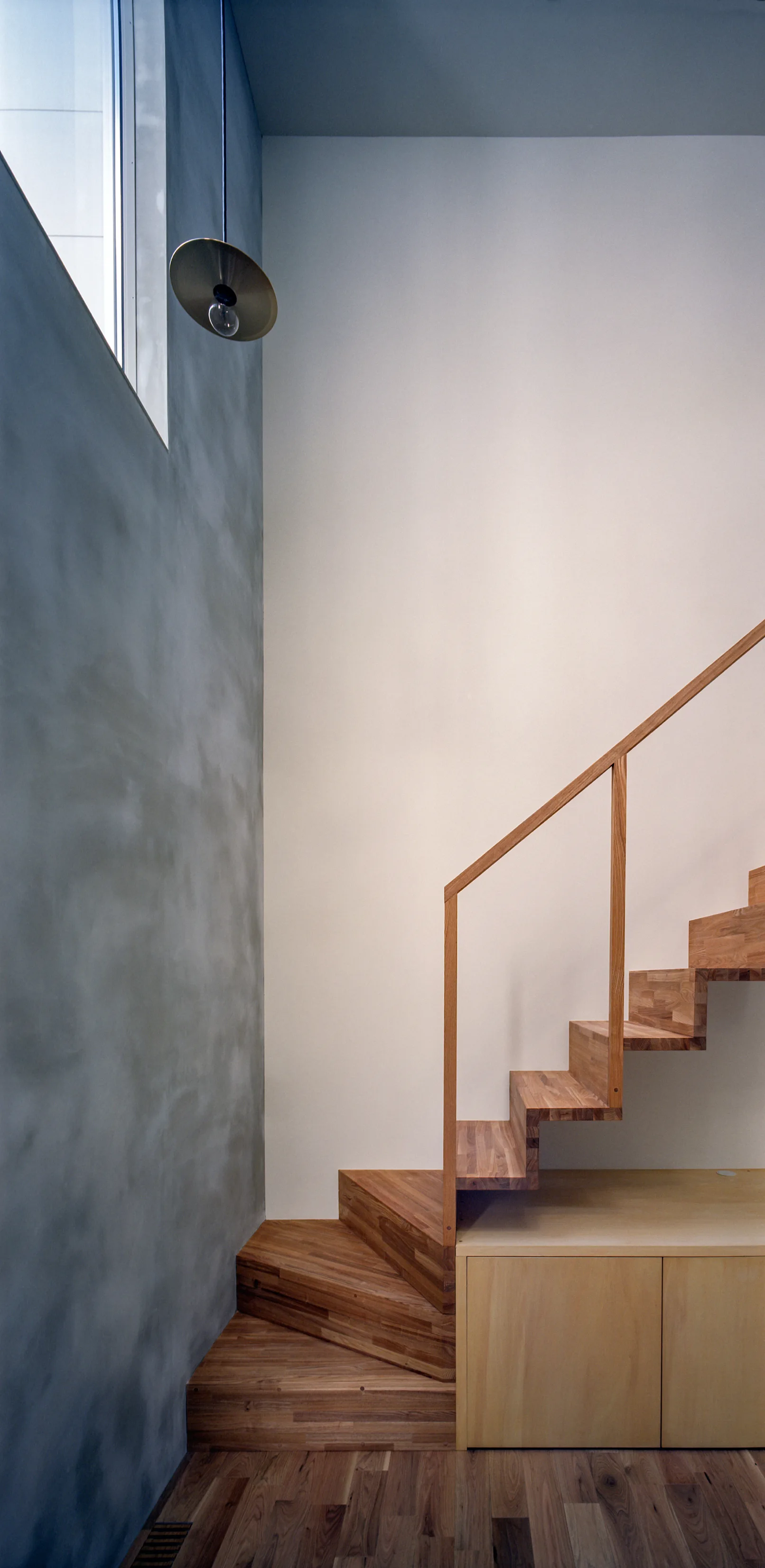 A wooden staircase with a simple wood handrail sits beside a textured grey plaster wall and features a light wood storage cabinet built underneath.